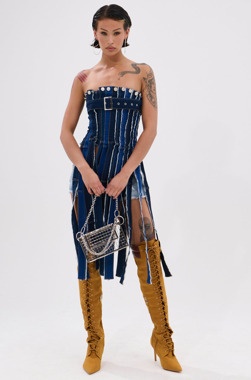 A person stands against a white background wearing the GOT A LOT GOING ON DENIM CORSET dress with vertical slits, paired with mustard yellow lace-up knee-high boots and holding a silver chain purse—a chic nod to upcycled fashion. 6