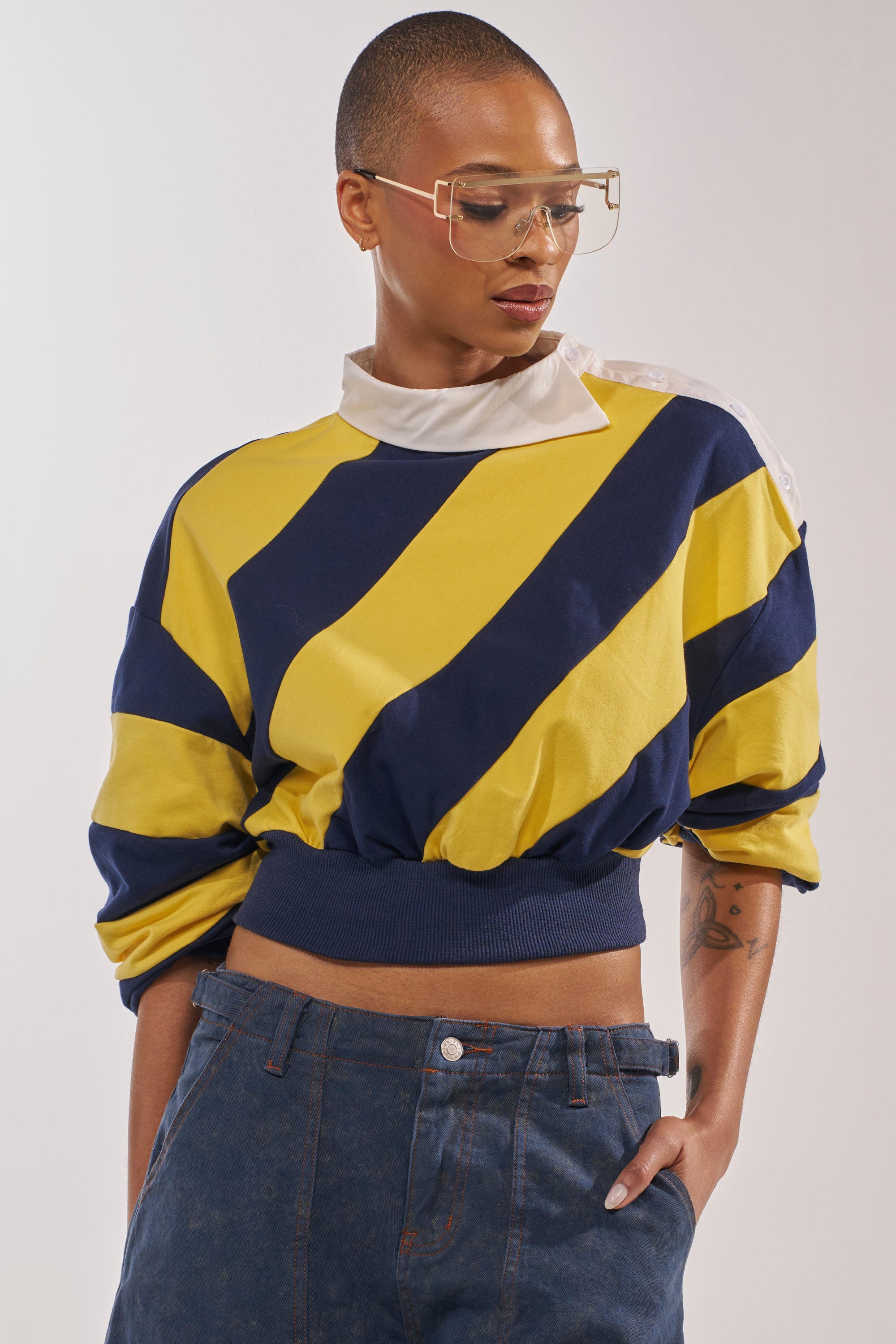 Wearing the JULIANA STRIPE SWEATSHIRT IN YELLOW, clear geometric glasses, and high-waisted denim jeans, a person stands with one hand in their pocket, looking down. 0