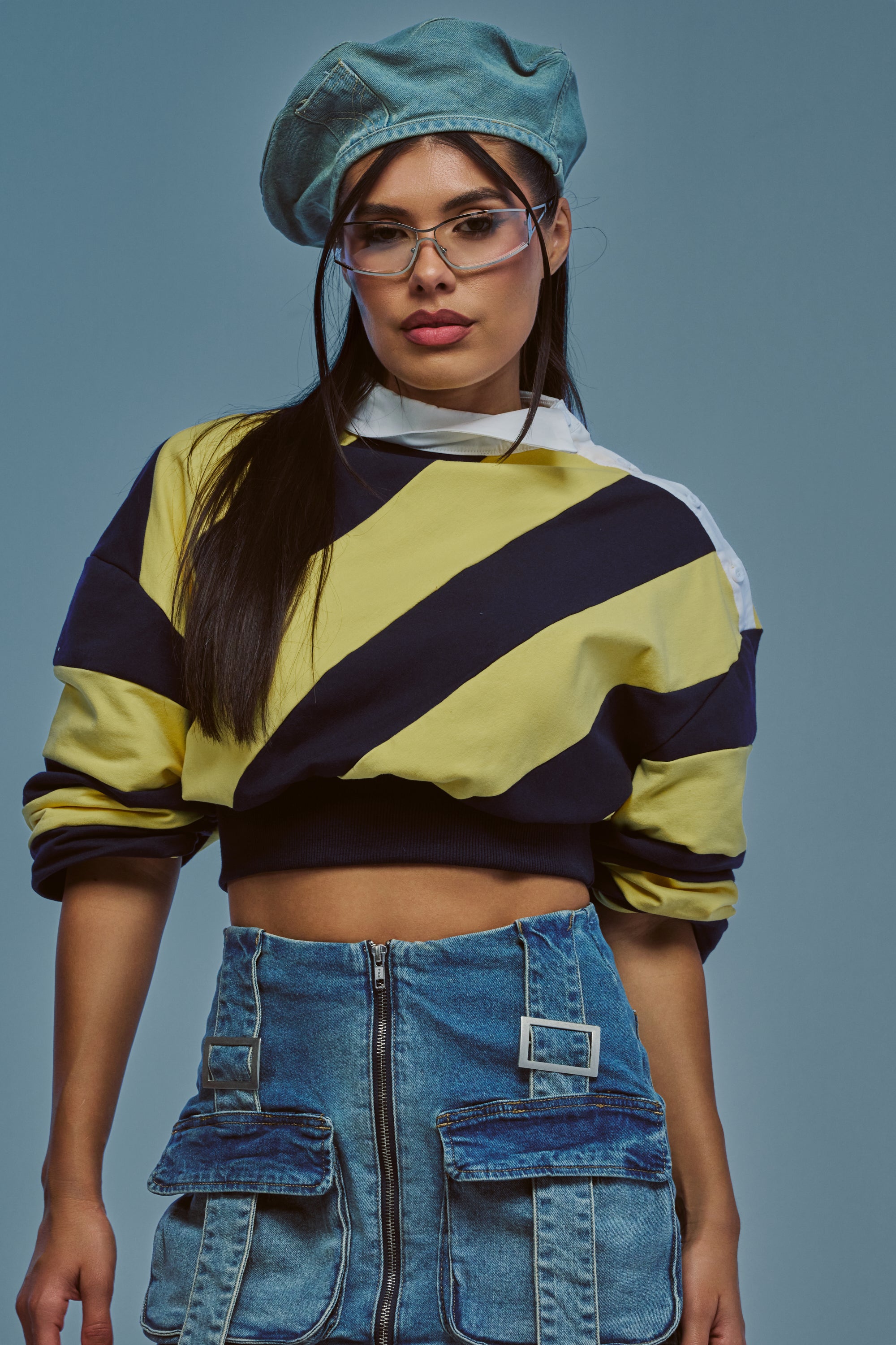 A woman wearing a beret, glasses, the JULIANA STRIPE SWEATSHIRT IN YELLOW, and a denim skirt with large pockets stands against a plain blue background.