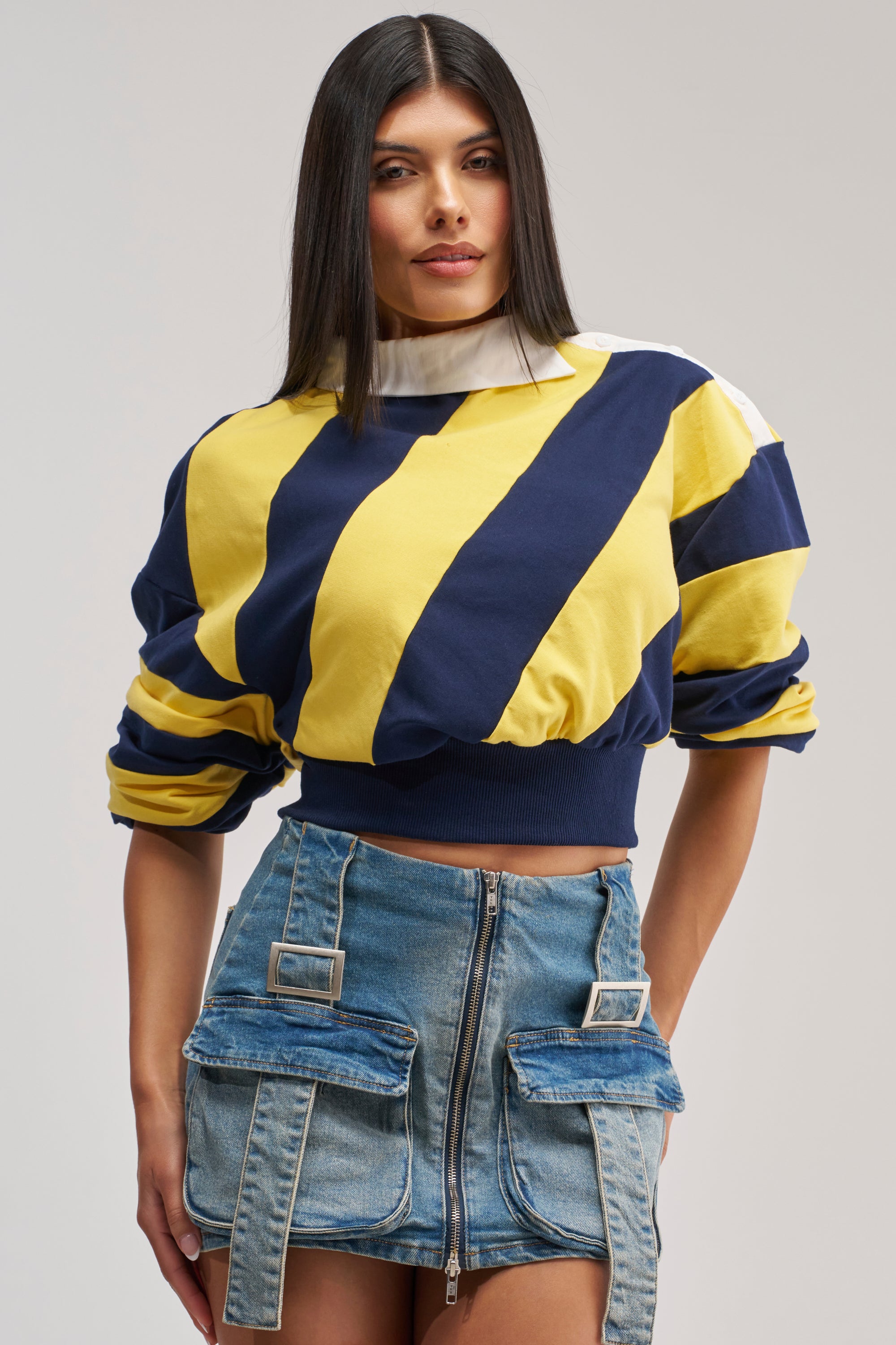 A woman with straight dark hair wears the JULIANA STRIPE SWEATSHIRT IN YELLOW, a yellow and navy cotton cropped striped shirt, styled with a blue denim mini skirt featuring prominent zipper and buckle accents, standing against a plain light gray background.