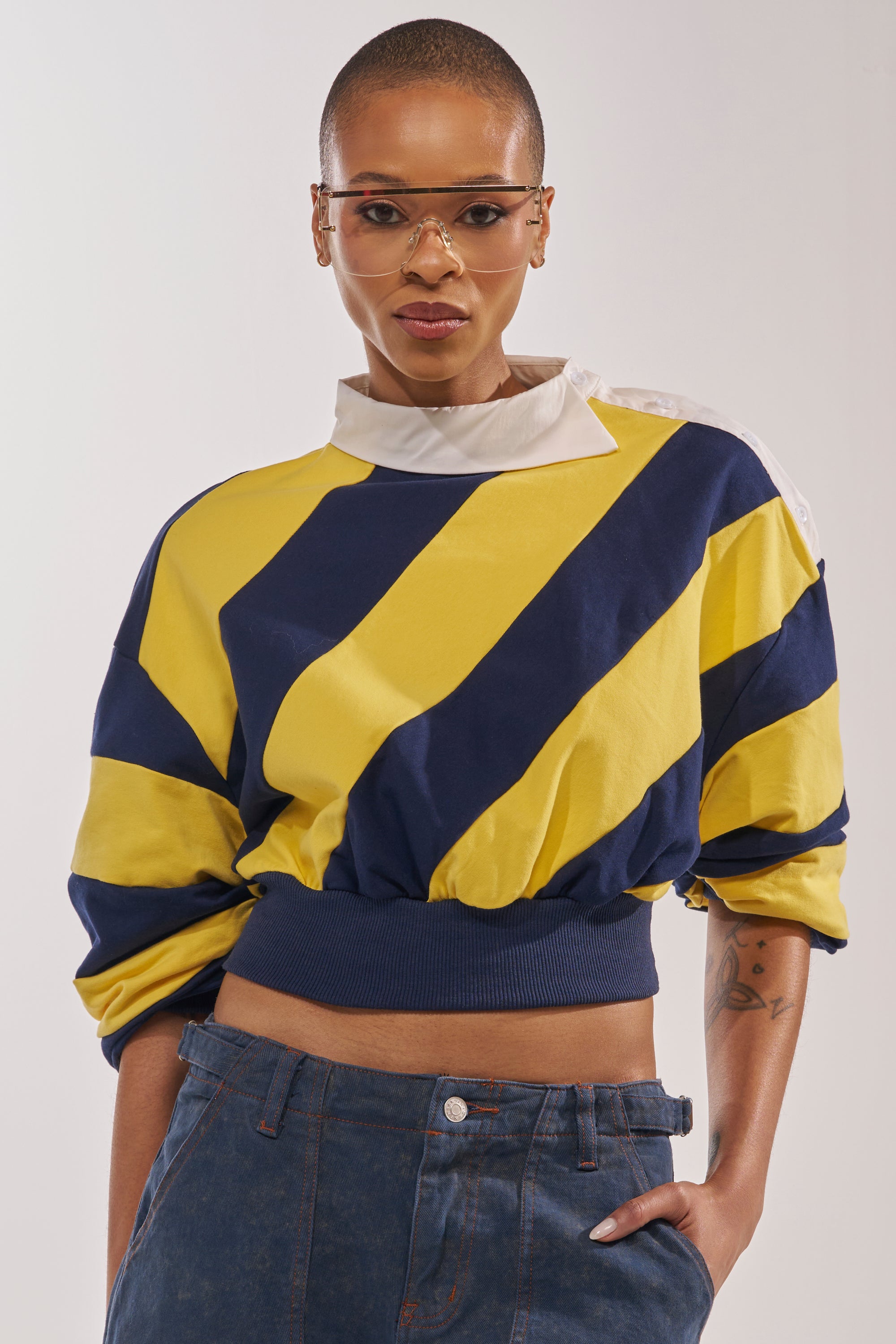 A person with short hair and sunglasses wears the JULIANA STRIPE SWEATSHIRT IN YELLOW, featuring an oversized collar and cropped fit, paired with blue jeans, standing confidently against a plain background. 1