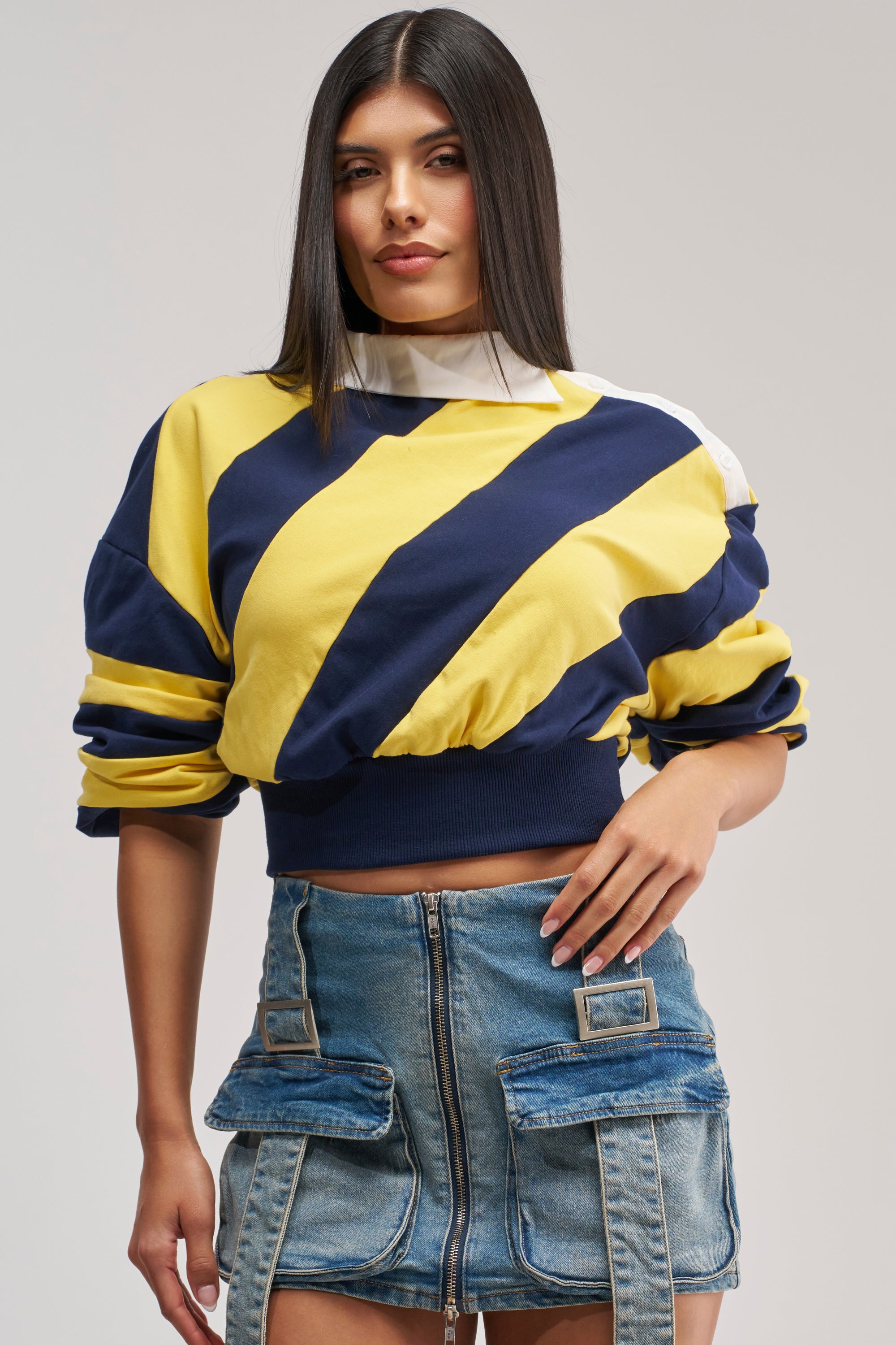 A woman with straight dark hair wears the Juliana Stripe Sweatshirt in Yellow—a cropped cotton shirt with navy and yellow stripes and an asymmetrical collar—styled with a denim skirt featuring a front zipper and utility pockets, standing against a neutral background.
