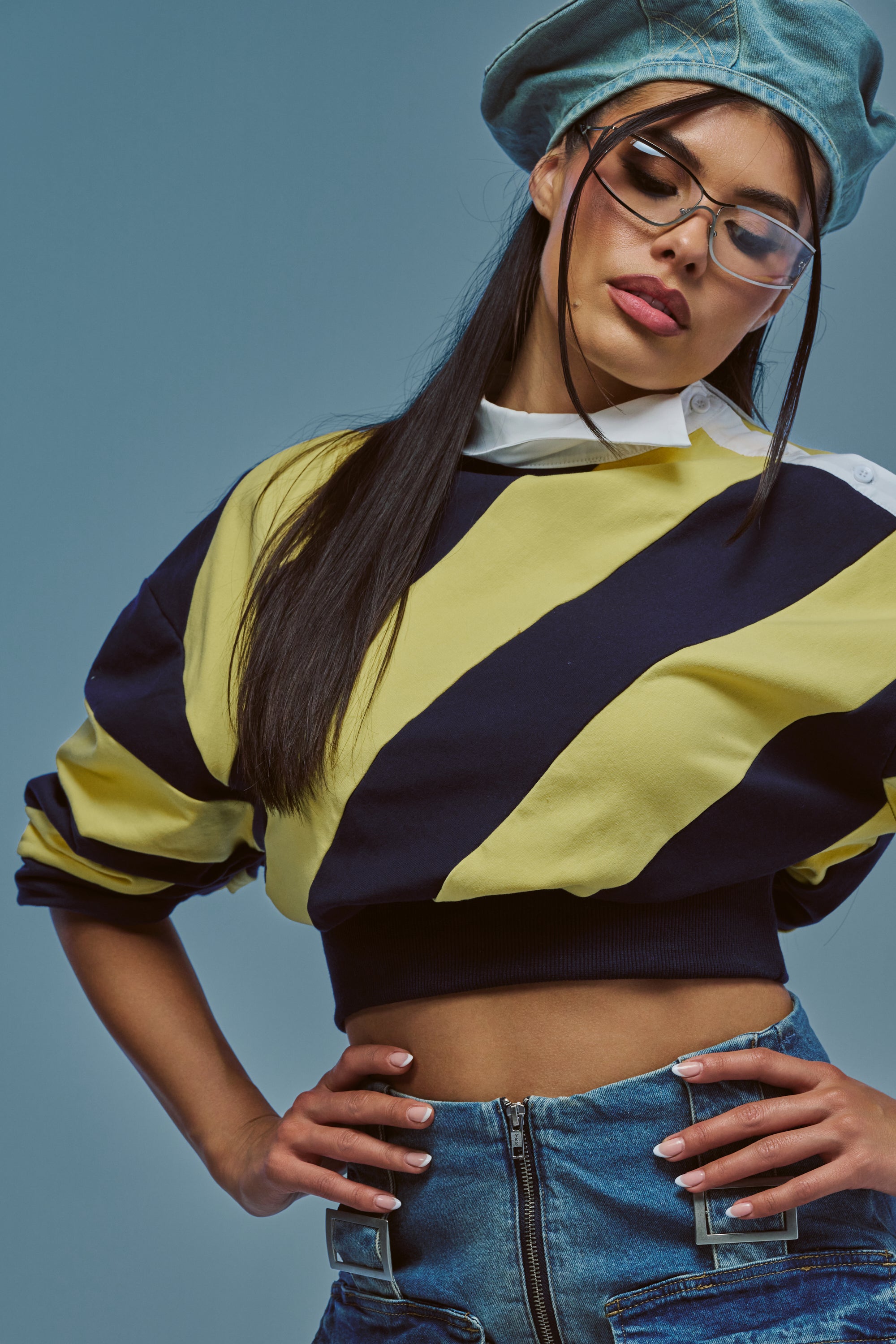 A woman wearing the JULIANA STRIPE SWEATSHIRT IN YELLOW, blue jeans, clear glasses, and a blue beret poses with her hands on her hips against a plain blue background.