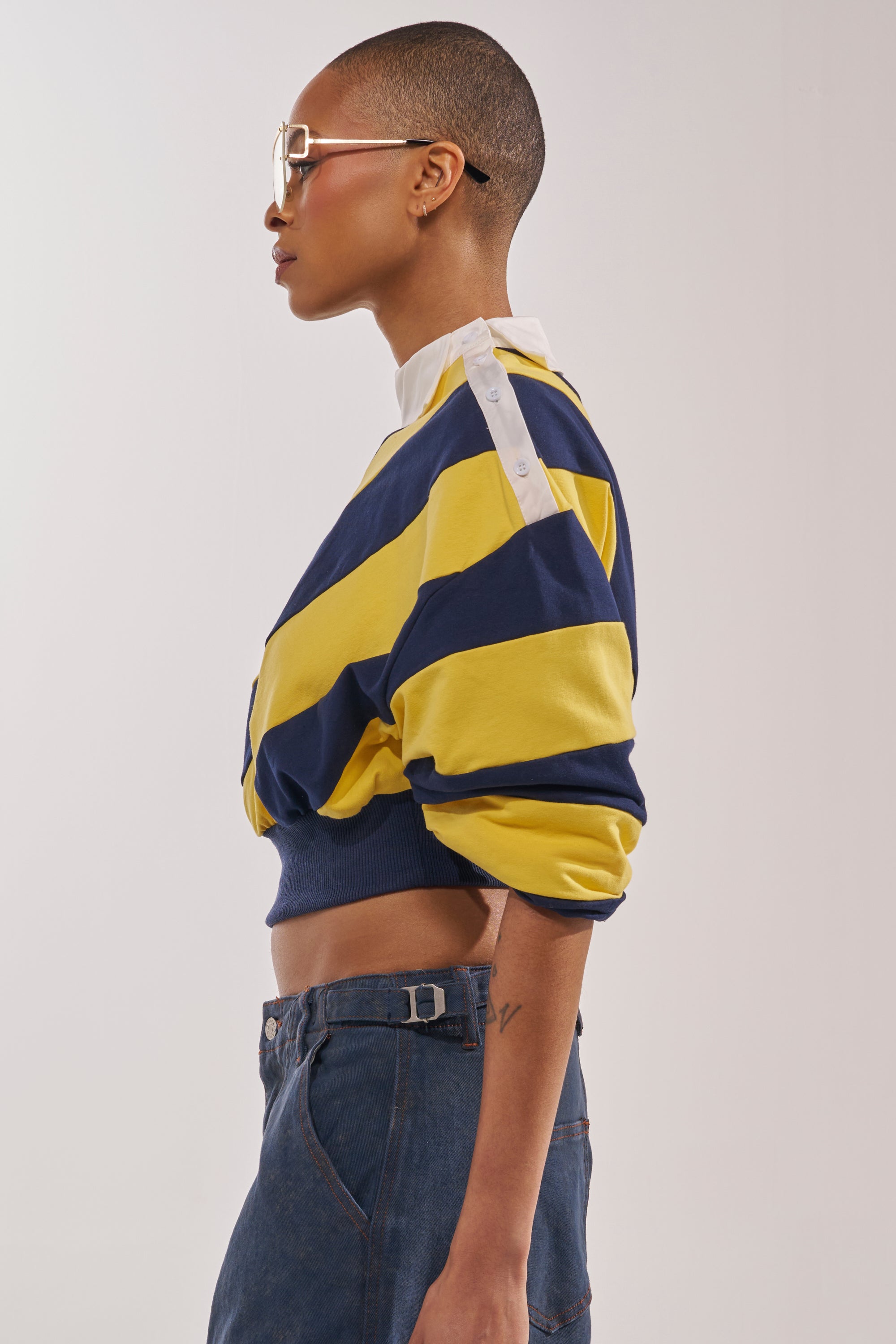 A person with a shaved head and large glasses wears the JULIANA STRIPE SWEATSHIRT IN YELLOW with an oversized collar over a white top and high-waisted blue jeans, shown in profile against a plain background. 2