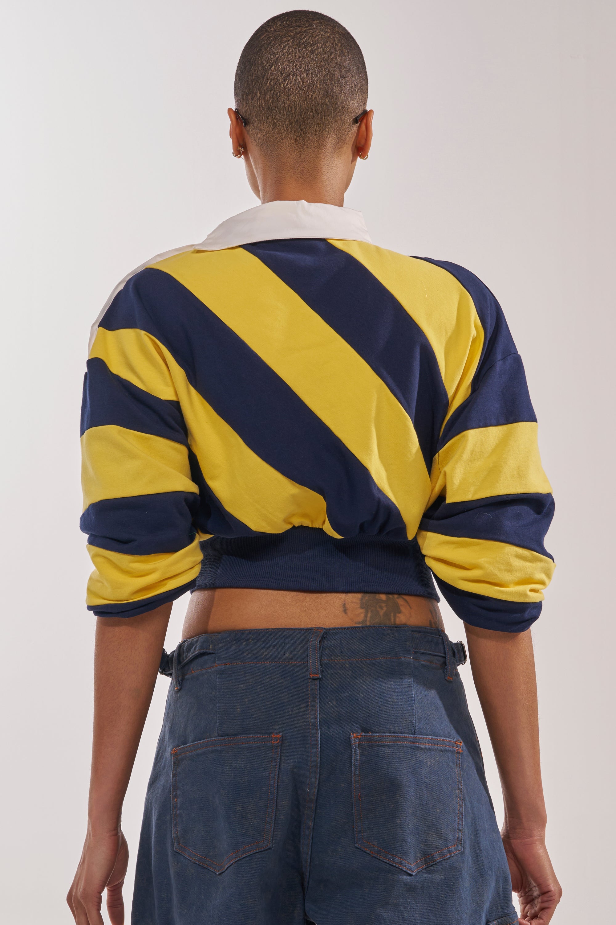 A person with a shaved head is seen from behind, wearing the JULIANA STRIPE SWEATSHIRT IN YELLOW—featuring bold yellow and navy stripes and an oversized collar—paired with loose blue jeans. Their arms rest relaxed at their sides. 3