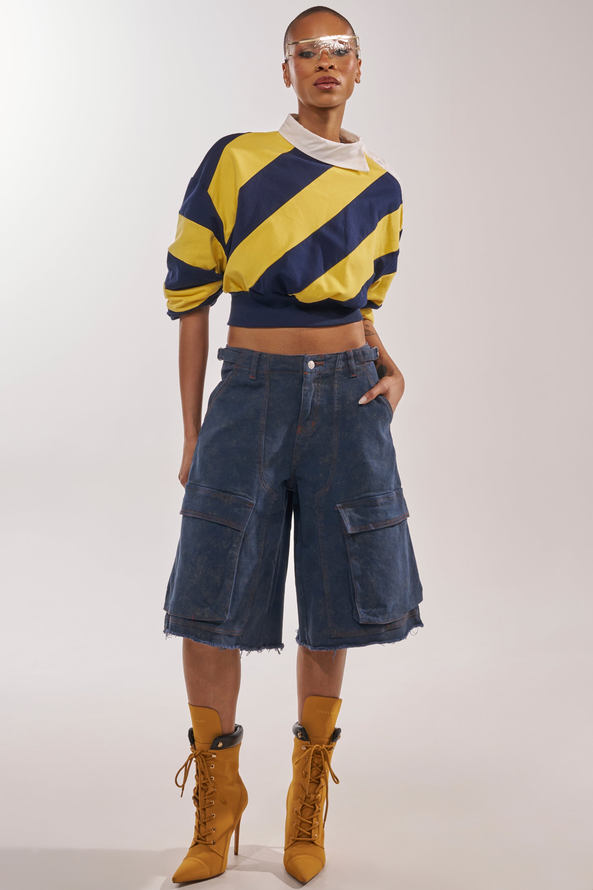 A person stands confidently in the JULIANA STRIPE SWEATSHIRT IN YELLOW, paired with loose knee-length denim cargo shorts, mustard lace-up boots, and clear visor glasses against a plain background. 4