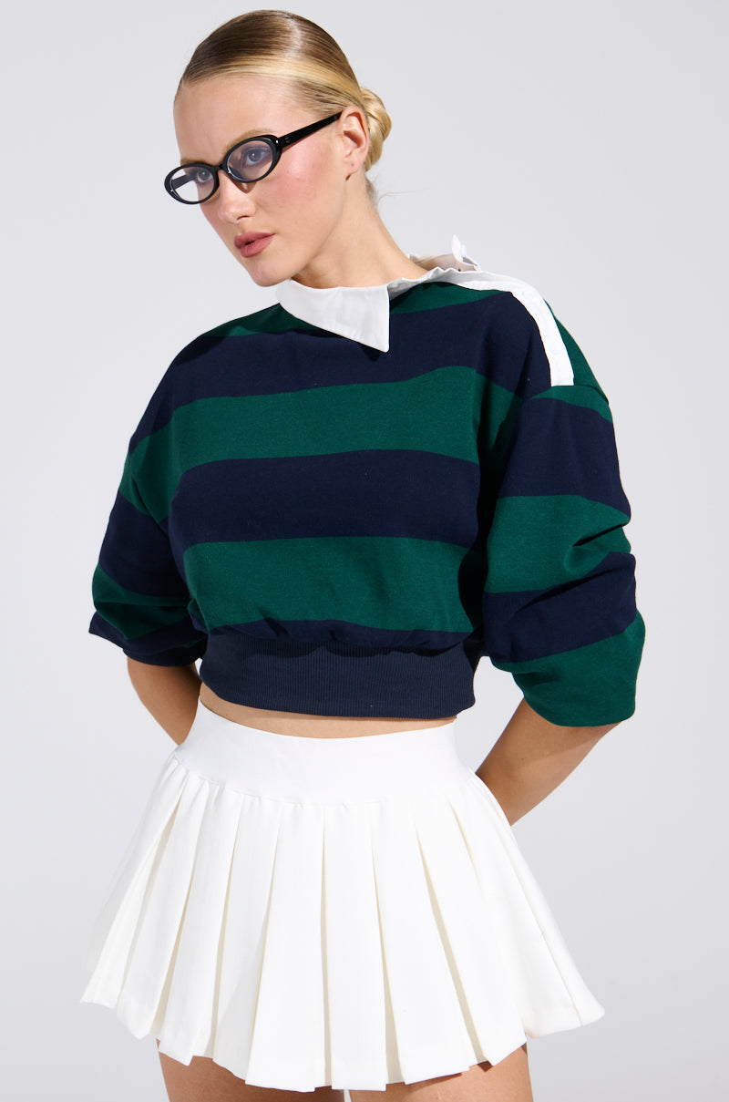 A woman in round black glasses, a JULIANA STRIPE SWEATSHIRT cropped over a white collared shirt, and a white pleated skirt stands against a plain light background.