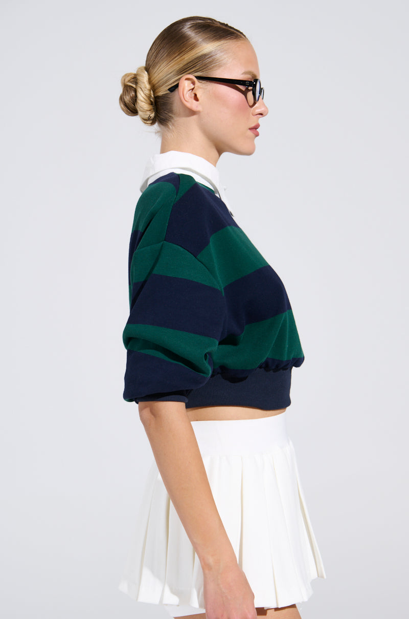 A woman with blonde hair in a neat bun and black glasses wears the JULIANA STRIPE SWEATSHIRT over a white collared shirt and a white pleated skirt, shown in profile against a plain background.