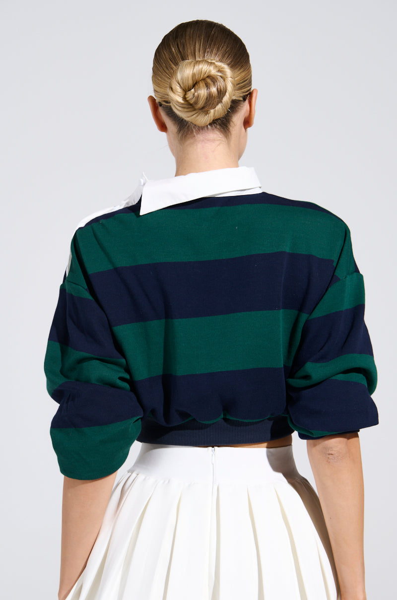 A person with blonde hair in a neat bun, wearing the JULIANA STRIPE SWEATSHIRT in green and navy with a white pleated skirt, is seen from the back against a plain light background.