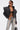 A woman with torso tattoos and slicked-back hair poses confidently in the COSMO ZIPPER MOTO jacket, black bra, ripped light-wash jeans, sunglasses, and a small handbag.