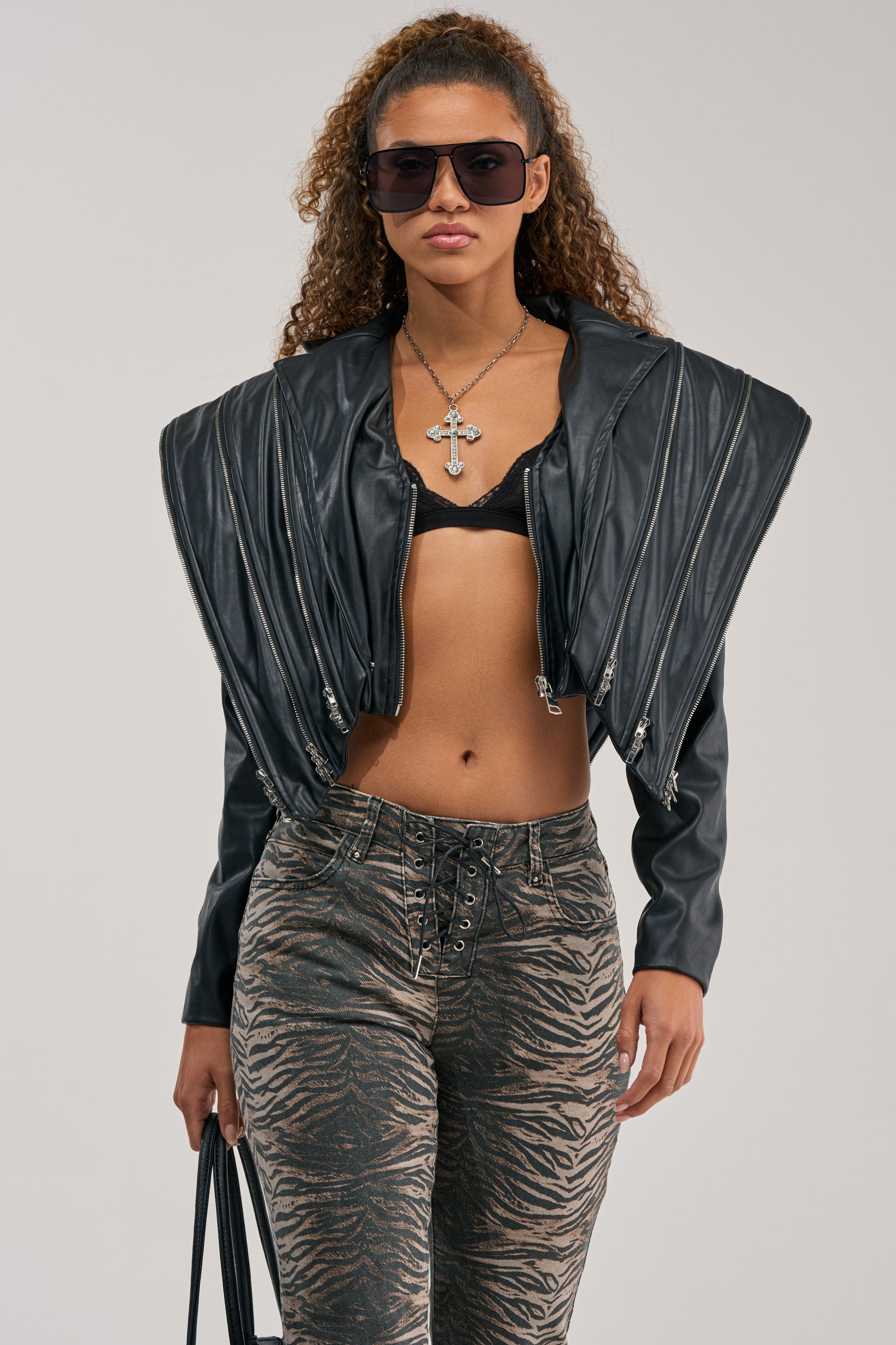 A woman with curly hair wears sunglasses, a black COSMO ZIPPER MOTO faux leather jacket with wide shoulders and an oversized collar, a black bralette, and tiger-print lace-up pants. She holds a black bag and wears a cross necklace.