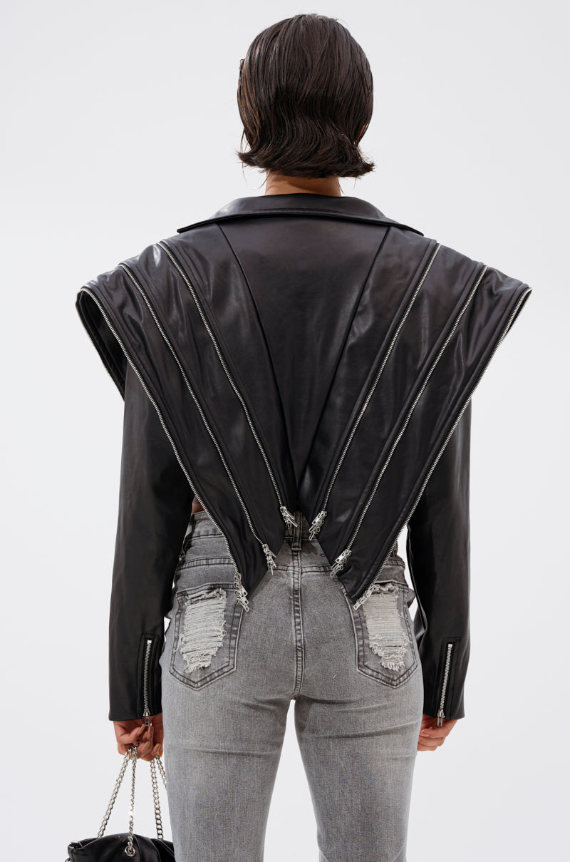 A person with short dark hair wears the COSMO ZIPPER MOTO—a faux leather jacket with dramatic pleats and exposed zippers—paired with light grey distressed jeans and carries a small chain-strap purse.
