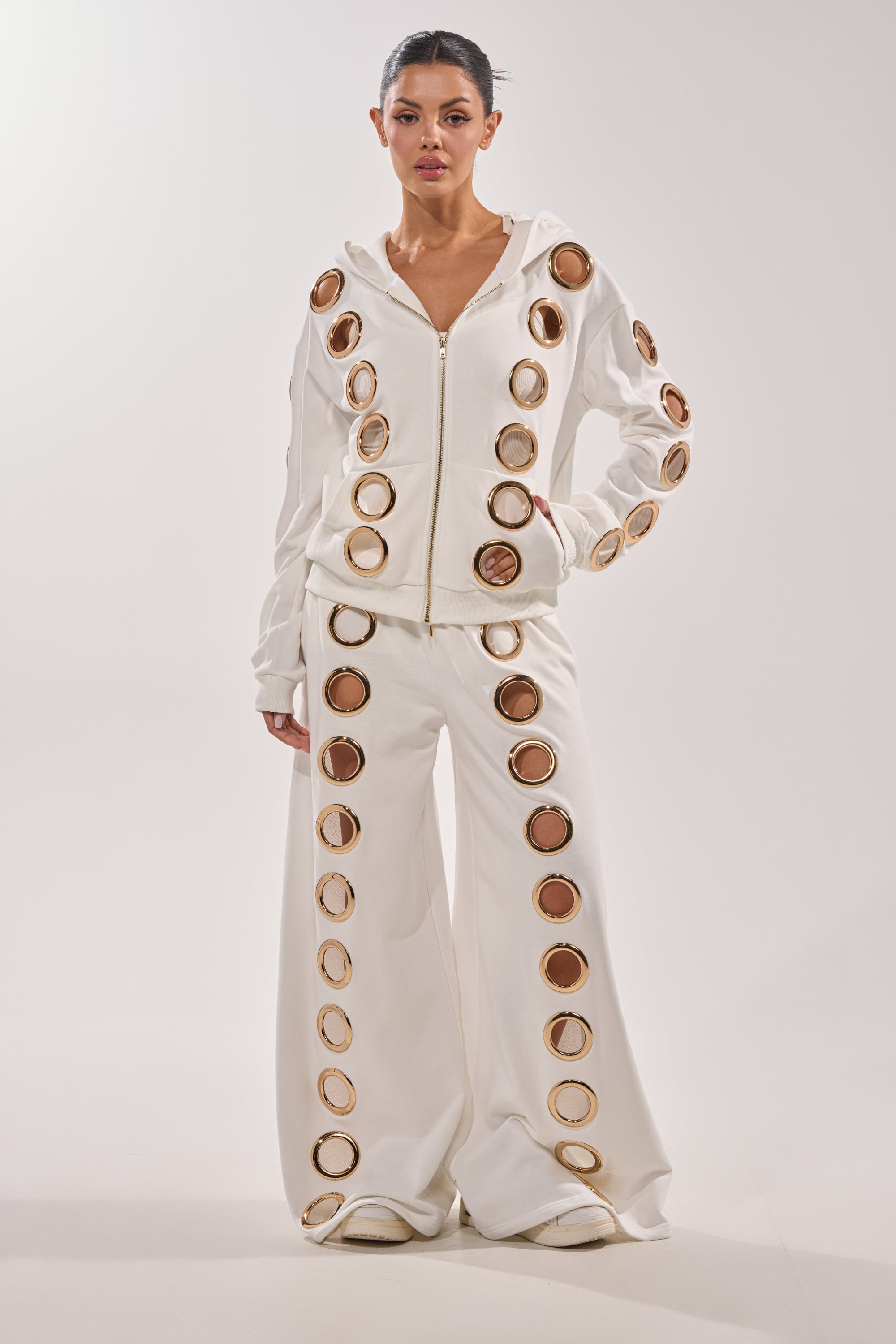 A woman poses in the THROUGH THE NIGHT WIDE LEG SWEATPANT WITH HOLES IN WHITE, paired with a luxe athleisure white hoodie, both featuring bold gold grommet ring details, against a plain light background. 0