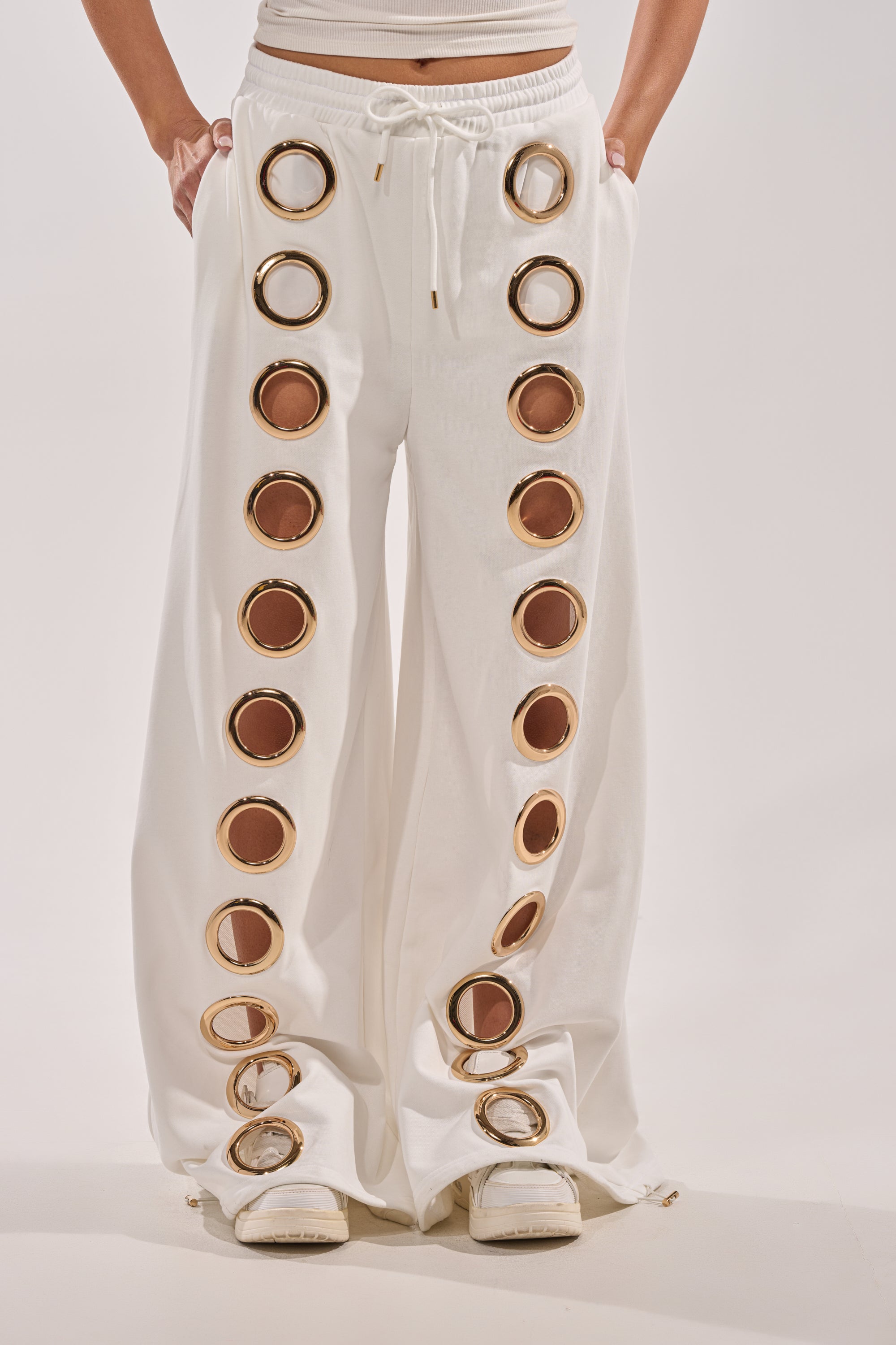 Person wearing THROUGH THE NIGHT WIDE LEG SWEATPANT WITH HOLES IN WHITE, featuring large circular cutouts down the sides, paired with white sandals and a cropped top, against a plain background. 1