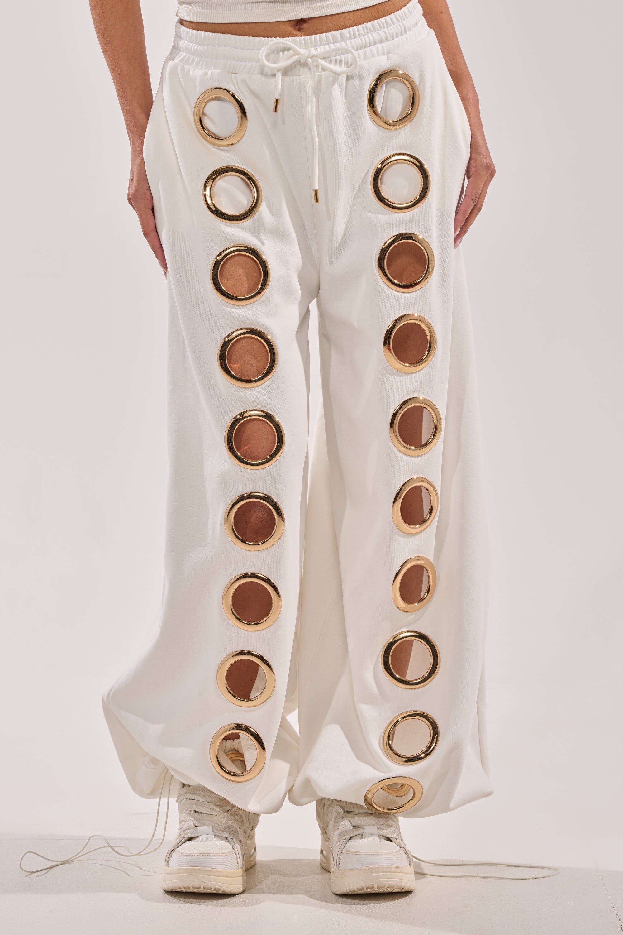 A person wearing the THROUGH THE NIGHT WIDE LEG SWEATPANT WITH HOLES IN WHITE, featuring two vertical rows of large gold-rimmed circular cutouts, paired with white sneakers and shown from waist to feet. 2