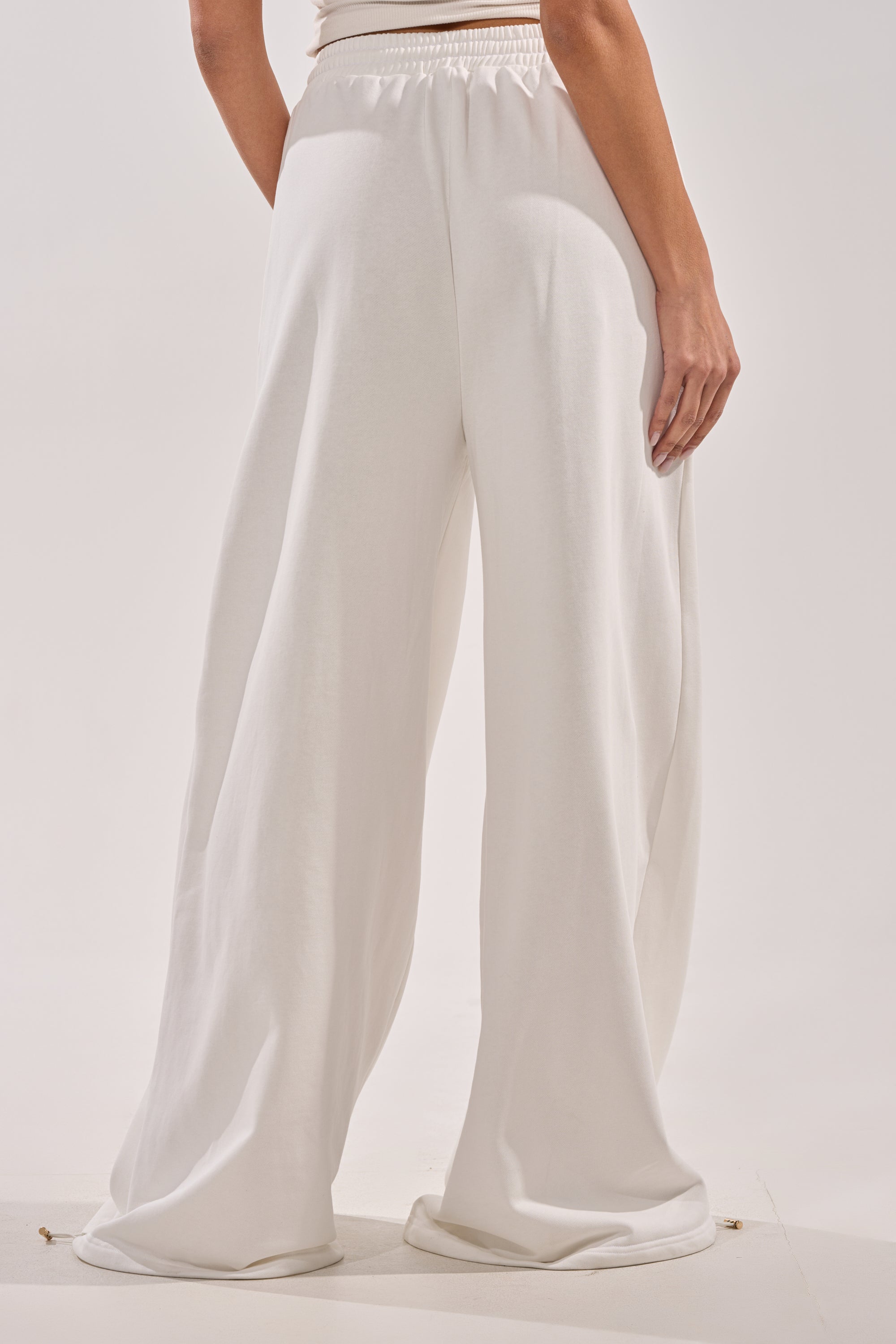 Someone wears the THROUGH THE NIGHT WIDE LEG SWEATPANT WITH HOLES IN WHITE, standing with their back to the camera to highlight its flowy fabric, elastic waistband, and laid-back athleisure style against a plain backdrop. 4