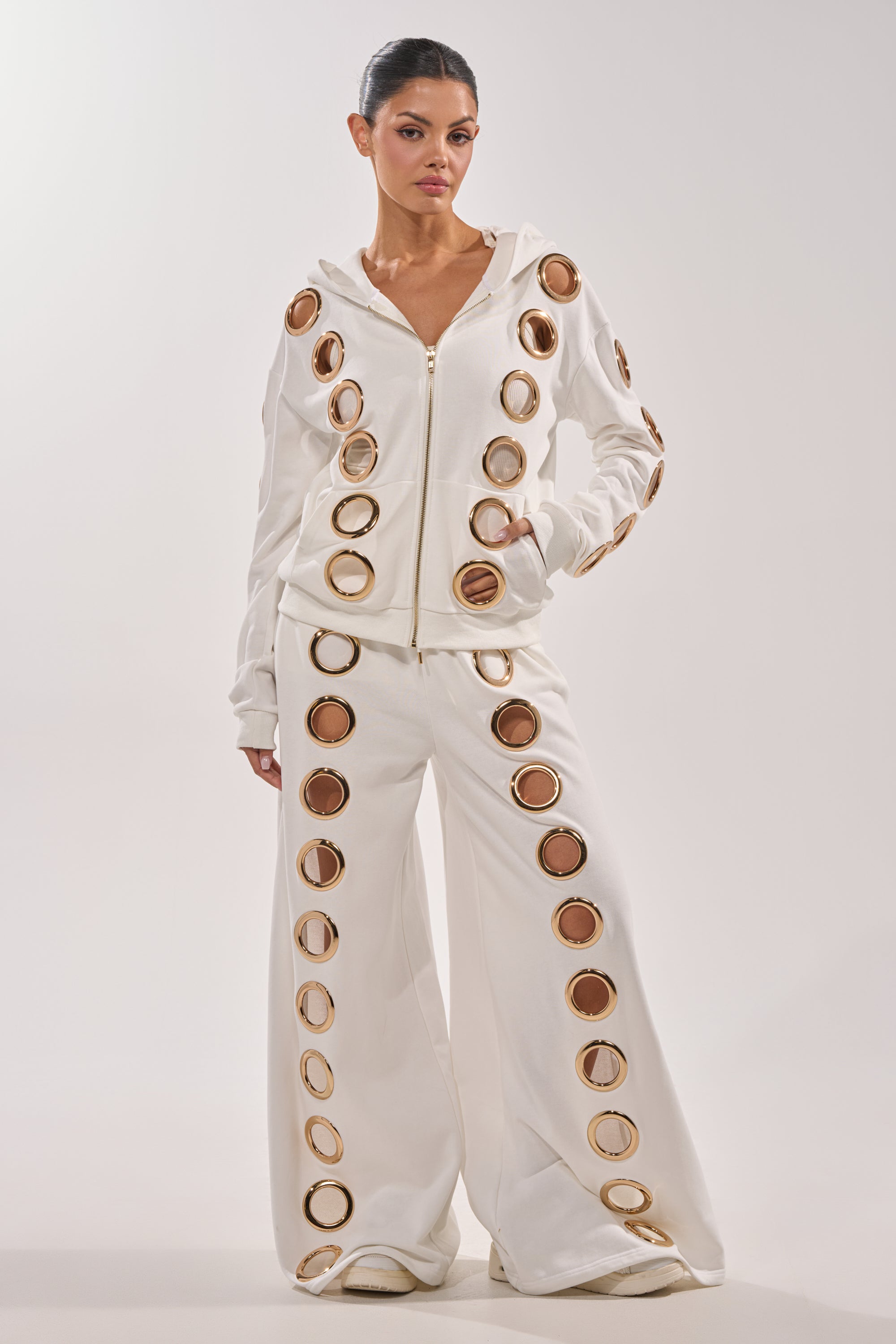 A woman stands against a plain light background in THROUGH THE NIGHT WIDE LEG SWEATPANT WITH HOLES IN WHITE, styled with a white zip-up hoodie and bold gold grommet rings for an athleisure look with striking metallic accents. 5