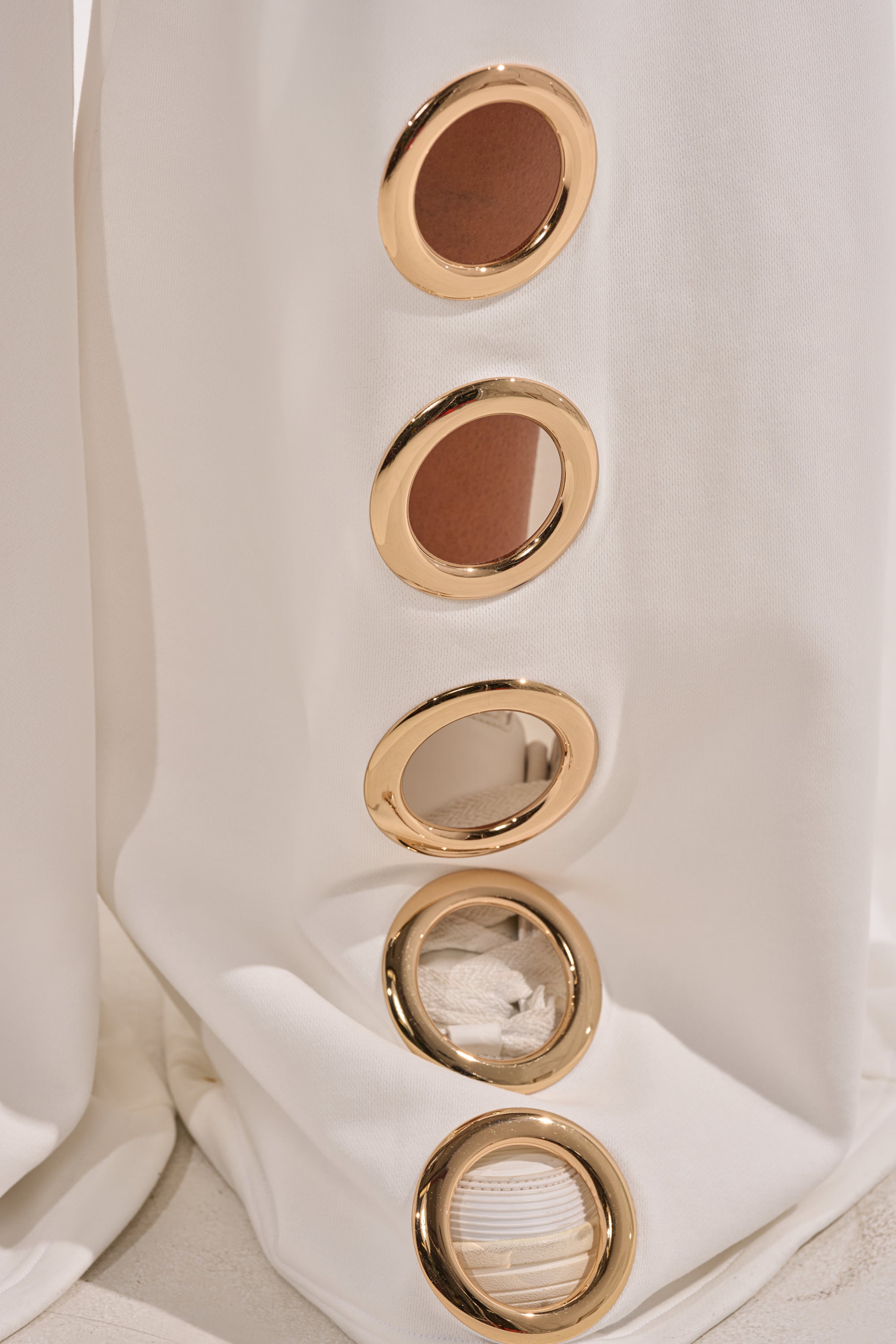 A close-up of the THROUGH THE NIGHT WIDE LEG SWEATPANT WITH HOLES IN WHITE shows five gold grommet rings arranged vertically in athleisure-style white fabric, each cutout displaying textured material and a neutral background. 6