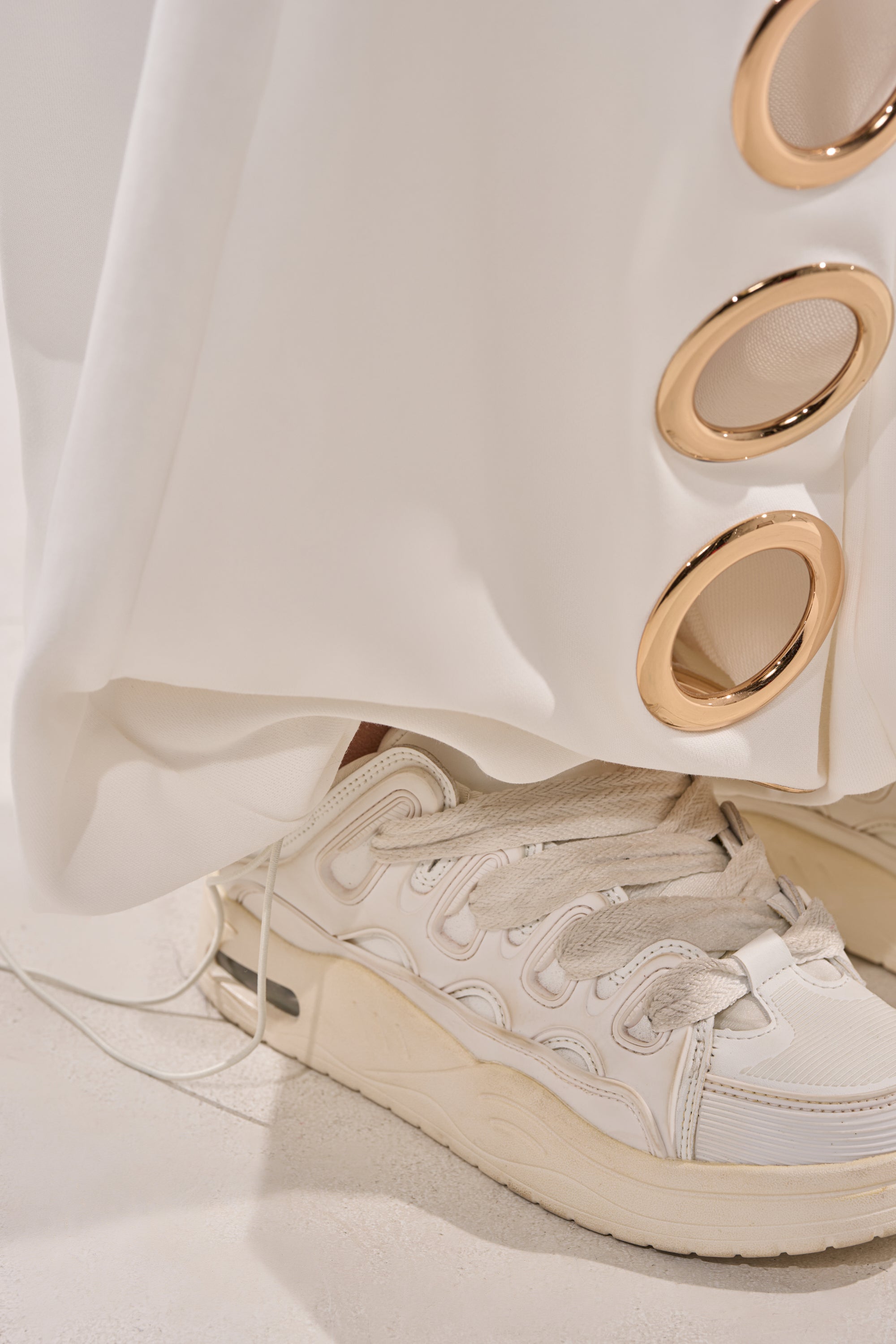 A close-up of the THROUGH THE NIGHT WIDE LEG SWEATPANT WITH HOLES IN WHITE paired with chunky white sneakers, showcasing athleisure style with standout metallic accents and bold grommet details. 7