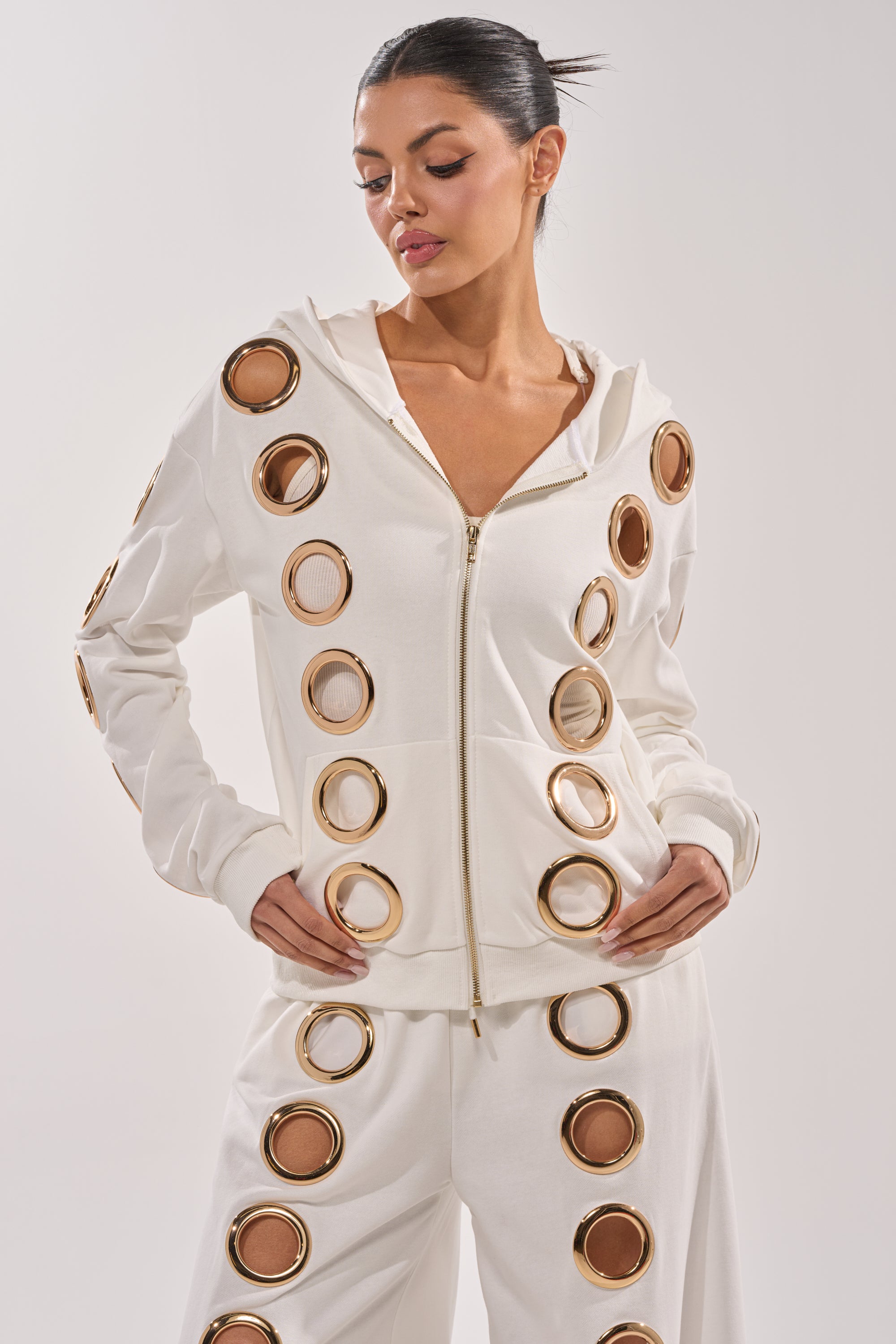 A woman models the THROUGH THE NIGHT SWEATSHIRT WITH HOLES IN WHITE, a luxe streetwear zip-up hoodie with bold gold grommet ring detailing, paired with matching pants. She stands hands on hips, looking down against a plain light background. 1