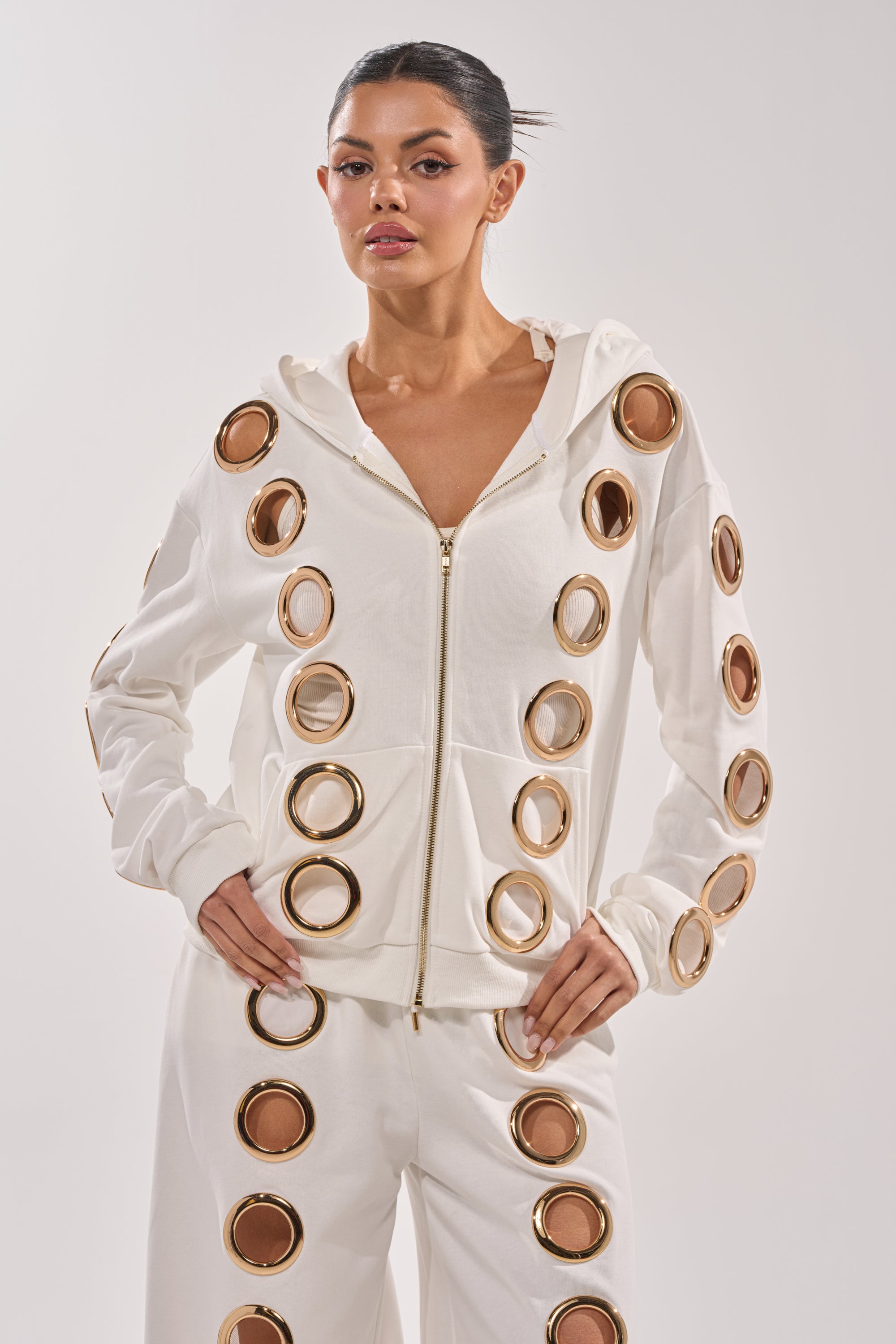 A woman wears the THROUGH THE NIGHT SWEATSHIRT WITH HOLES IN WHITE, a luxe zip-up hoodie with metallic gold grommet rings and front pockets, styled with matching pants against a neutral background. 2