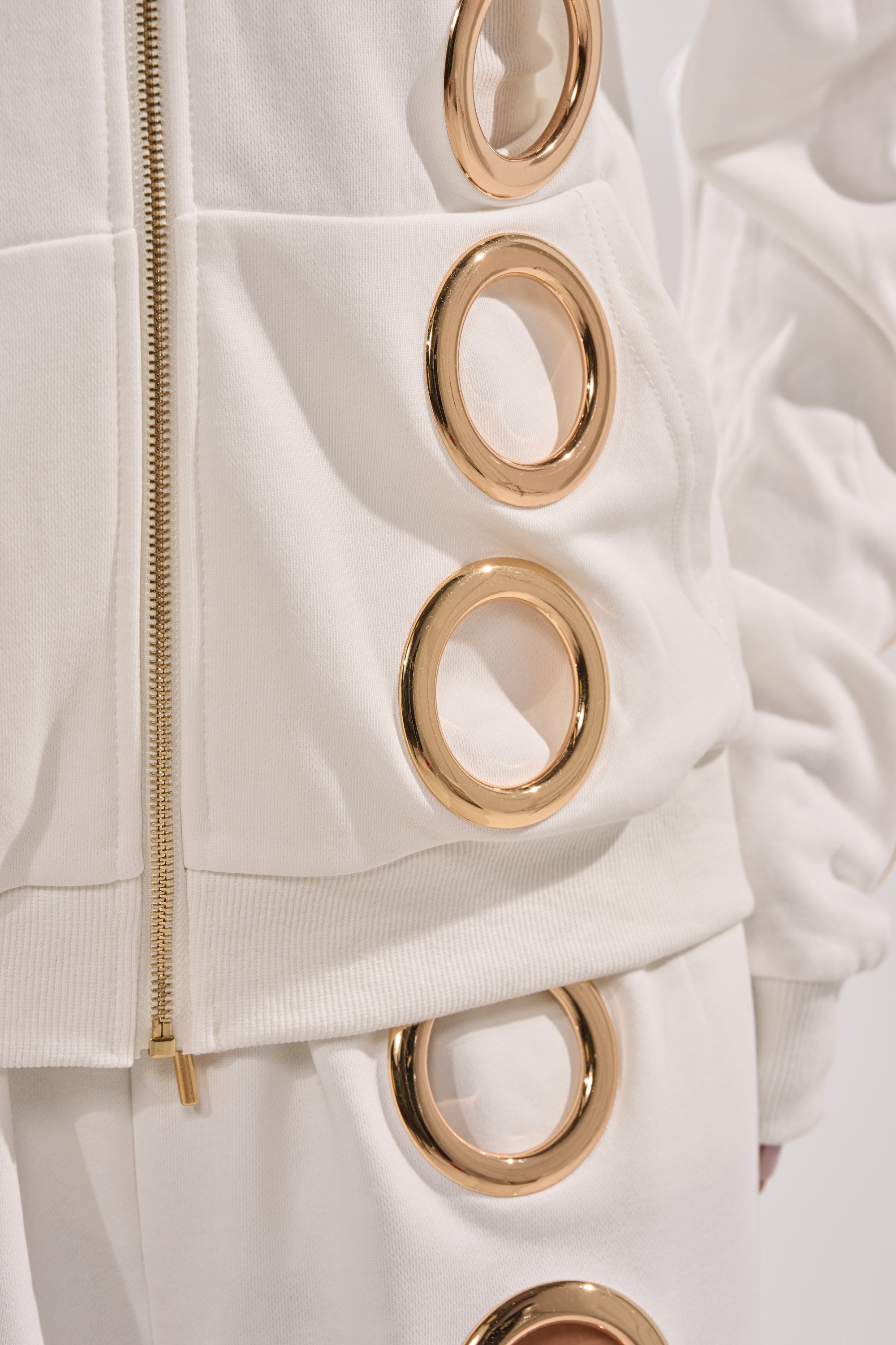 Close-up of a luxe streetwear look featuring the THROUGH THE NIGHT SWEATSHIRT WITH HOLES IN WHITE, detailed with large gold grommet rings and a striking gold zipper, paired with matching pants. 7