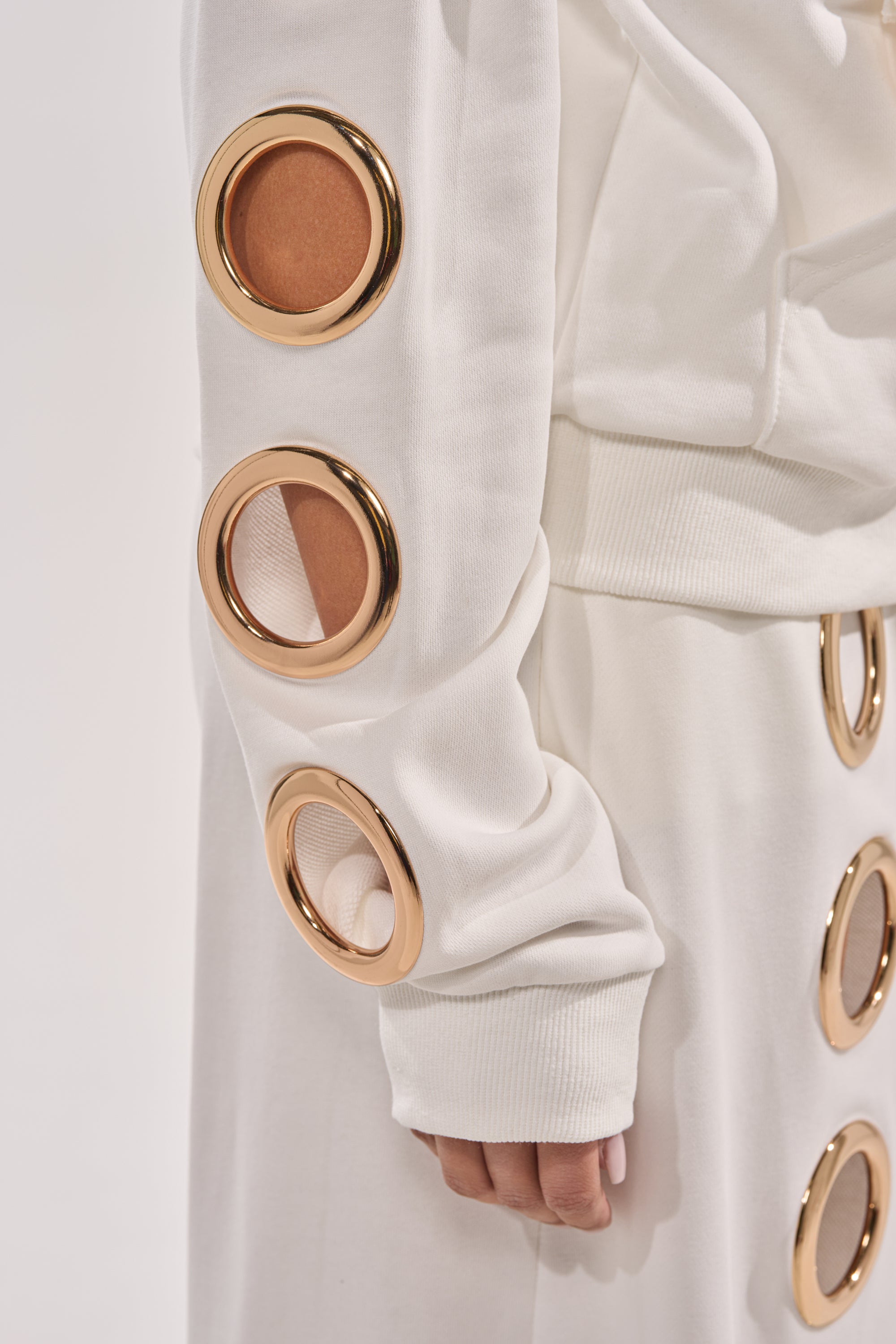 Wearing the THROUGH THE NIGHT SWEATSHIRT WITH HOLES IN WHITE, this luxe streetwear look features gold grommet ring details on the sleeves, with cutouts revealing glimpses of skin and fabric from mid-torso to hand. 8
