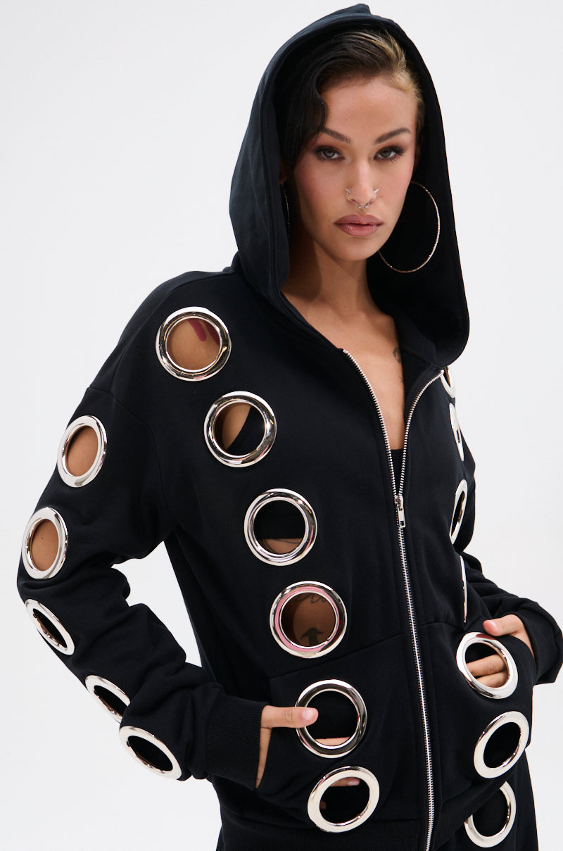 A woman poses in the THROUGH THE NIGHT SWEATSHIRT WITH HOLES, a black zip-up hoodie featuring large metallic grommet holes along the sleeves and front. She accessorizes with hoop earrings, a nose ring, and wears her hood up against a plain white background.