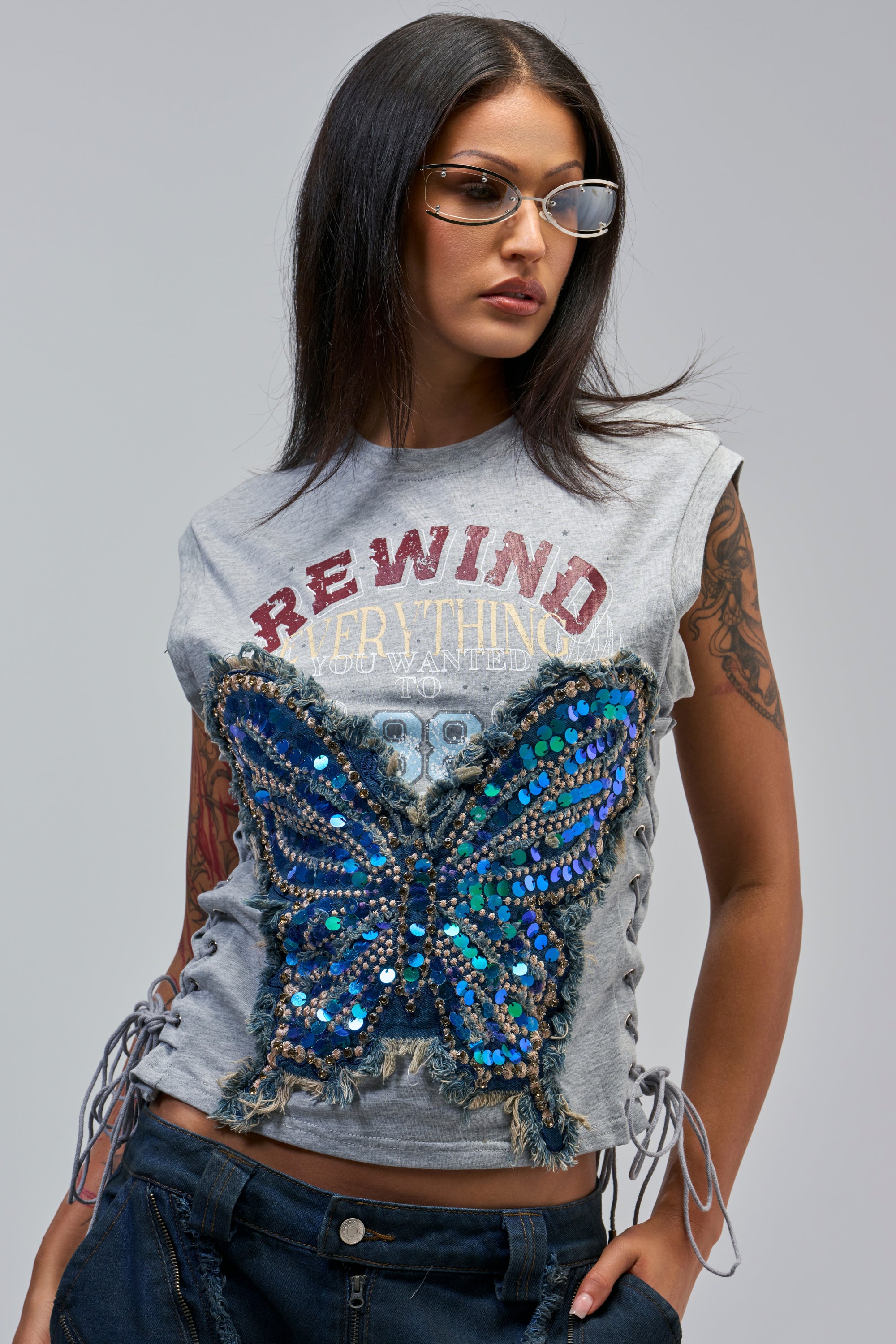 A woman with straight dark hair and tattoos wears sunglasses and the REWIND EVERYTHING TEE IN LIGHT GREY, styled with dark blue jeans for a festival-ready look.