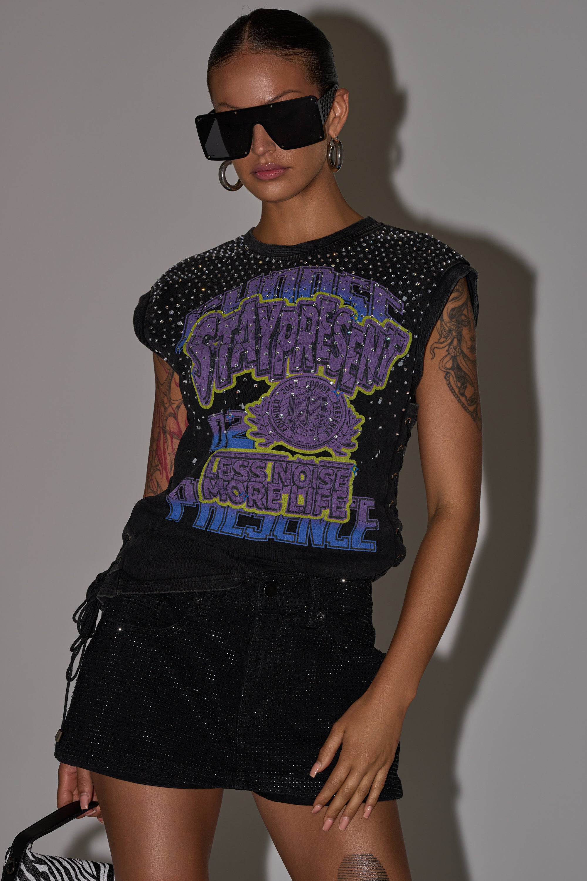 A woman confidently poses in the STAY PRESENT EMBELLISHED LACE UP TEE, styled with oversized black sunglasses, hoop earrings, and a studded mini skirt for a bold street-style look against a plain background. 0