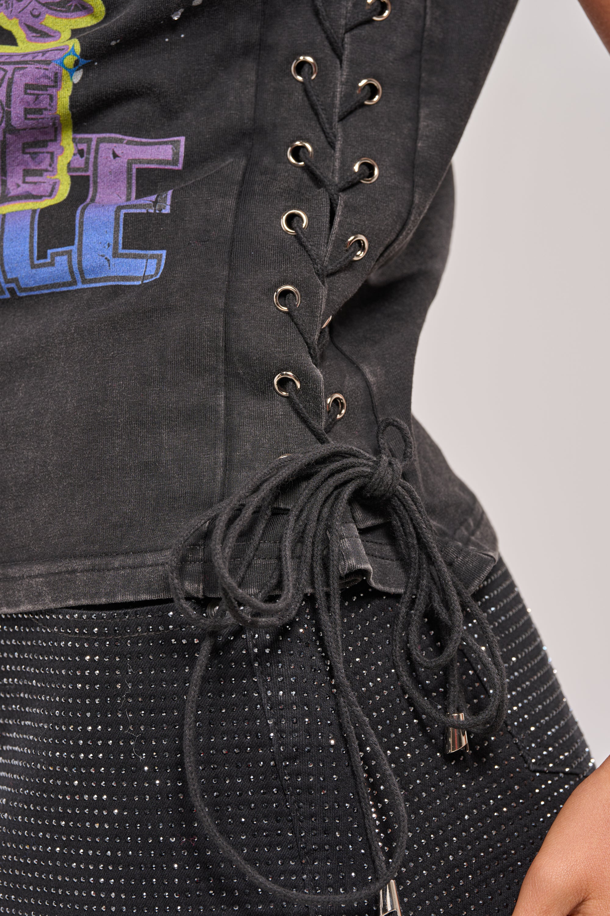 Close-up of a person wearing the STAY PRESENT EMBELLISHED LACE UP TEE with metal eyelets and a graphic print, paired with black pants featuring white studs. The tee's waist laces are tied in a bow, completing this edgy look. 9