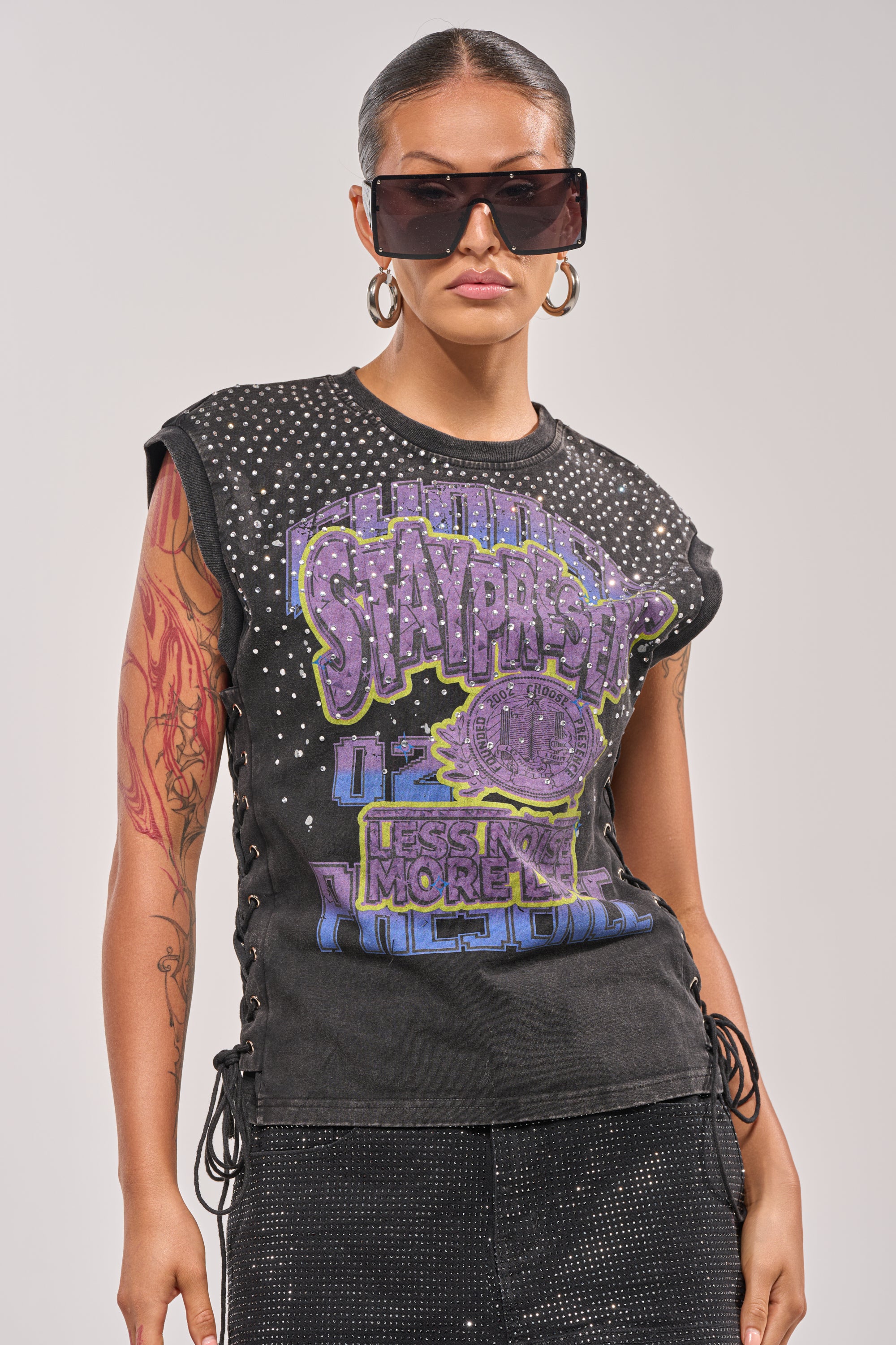 A woman with slicked-back hair, hoop earrings, and oversized sunglasses wears the STAY PRESENT EMBELLISHED LACE UP TEE featuring colorful text and rhinestones. She has a tattoo sleeve on her left arm and stands against a plain background. 1