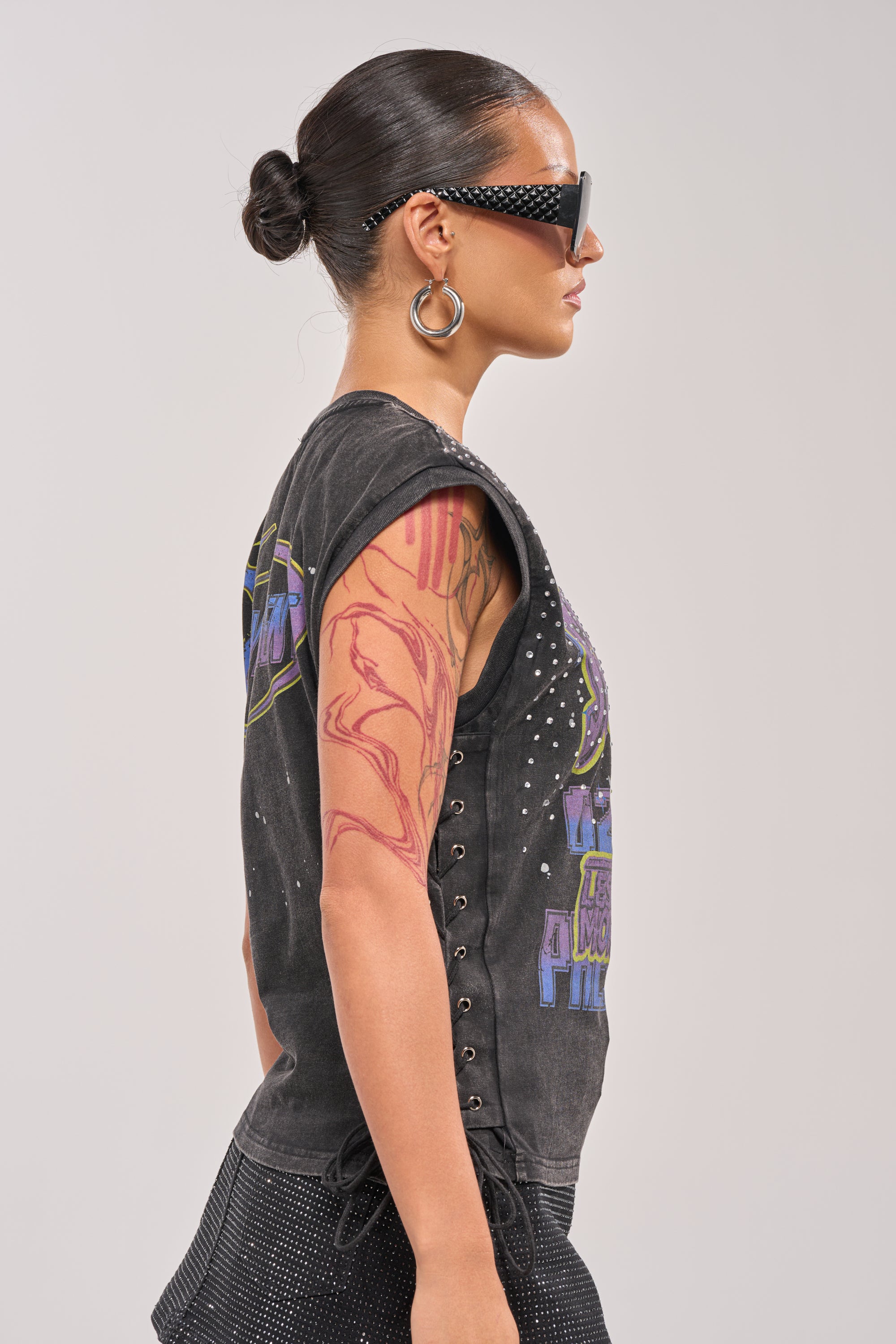 Woman in profile wears sunglasses, hoop earrings, and the STAY PRESENT EMBELLISHED LACE UP TEE—a black sleeveless top with side lacing. Her hair is in a sleek bun, she has an upper arm tattoo, and stands against a plain light grey background. 3
