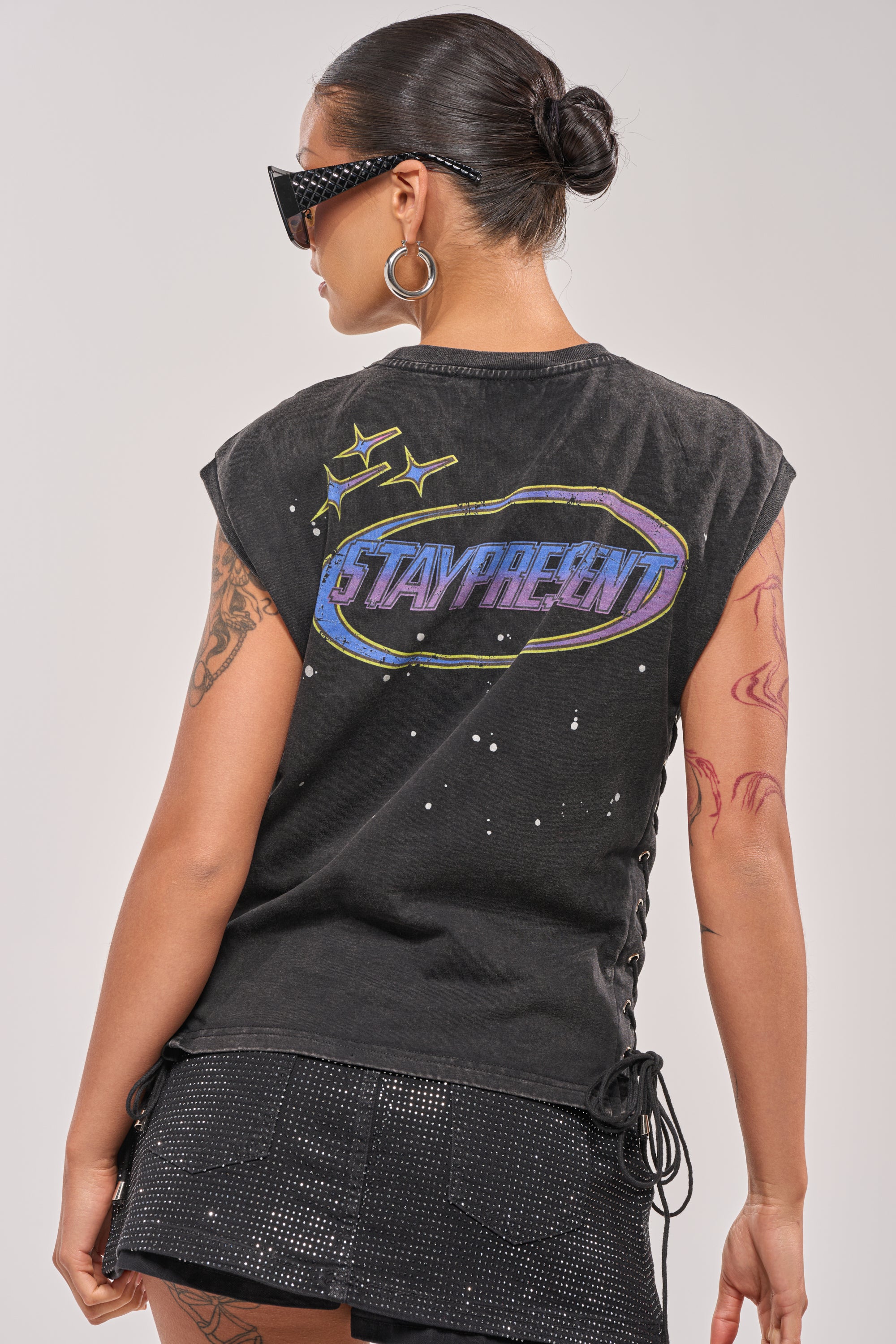 A woman with a bun, sunglasses, hoop earrings, and visible arm tattoos wears the STAY PRESENT EMBELLISHED LACE UP TEE—with colorful "STAY PRESENT" on the back—and a black skirt. 4