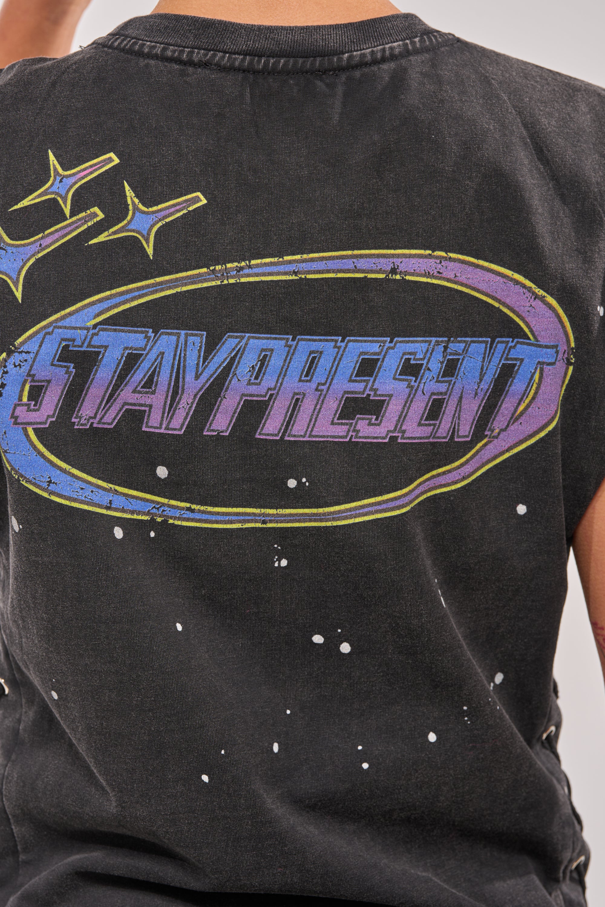 Someone wears the STAY PRESENT EMBELLISHED LACE UP TEE, a black top featuring "STAY PRESENT" in bold, colorful letters on the back, set inside a blue and yellow oval with star accents. 8