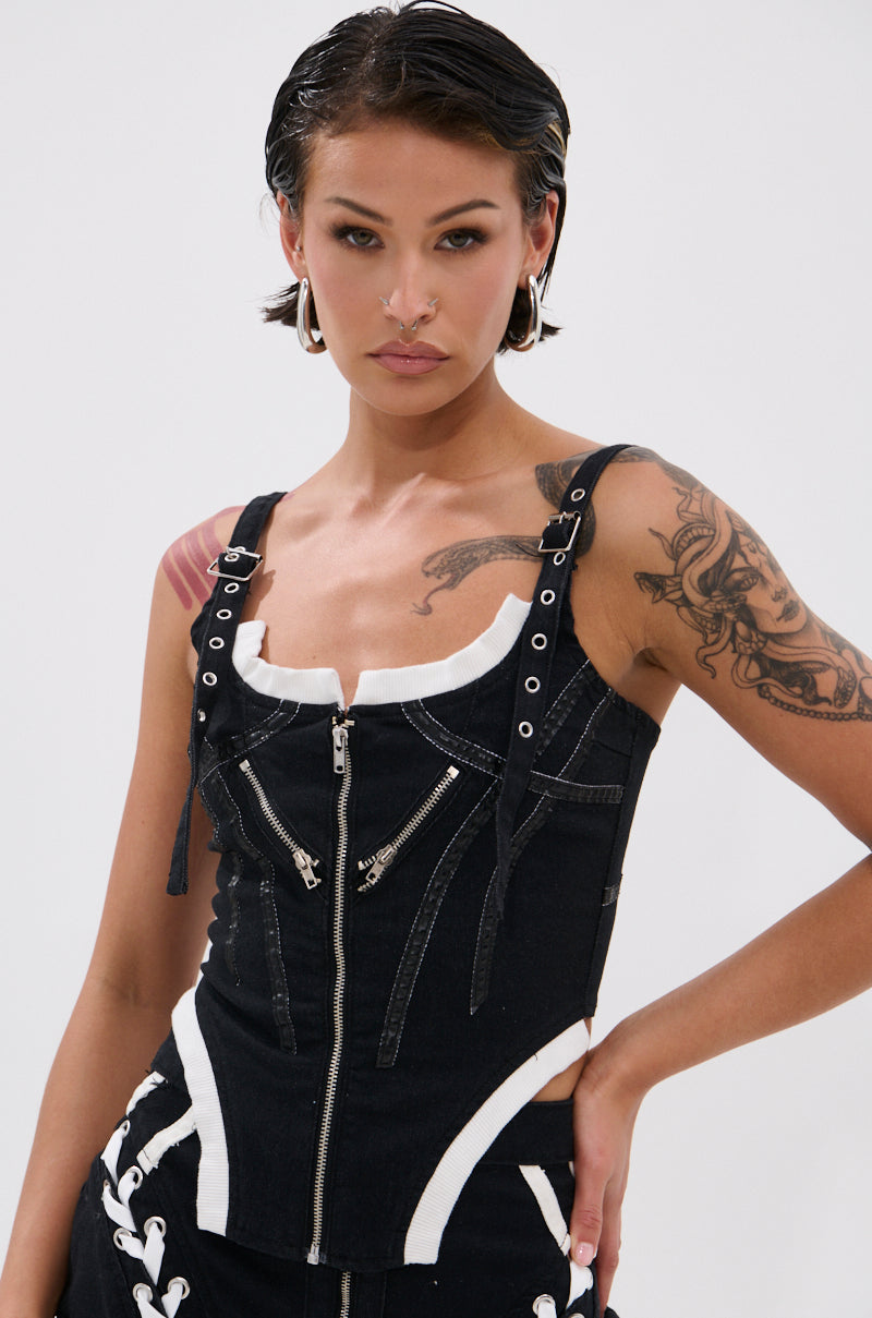 A woman with short dark slicked-back hair and arm tattoos wears the WRAPPED UP DENIM CORSET—a black and white top with faux leather, zipper, and strap accents—against a plain light background.