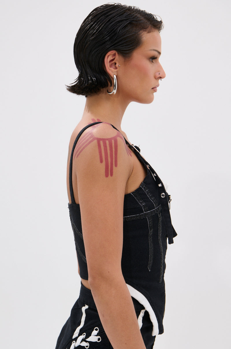 A person with short, slicked-back dark hair and hoop earrings stands in profile wearing the WRAPPED UP DENIM CORSET, featuring white stitching and faux leather accents. A painted red drip design highlights their shoulder against a plain white background.