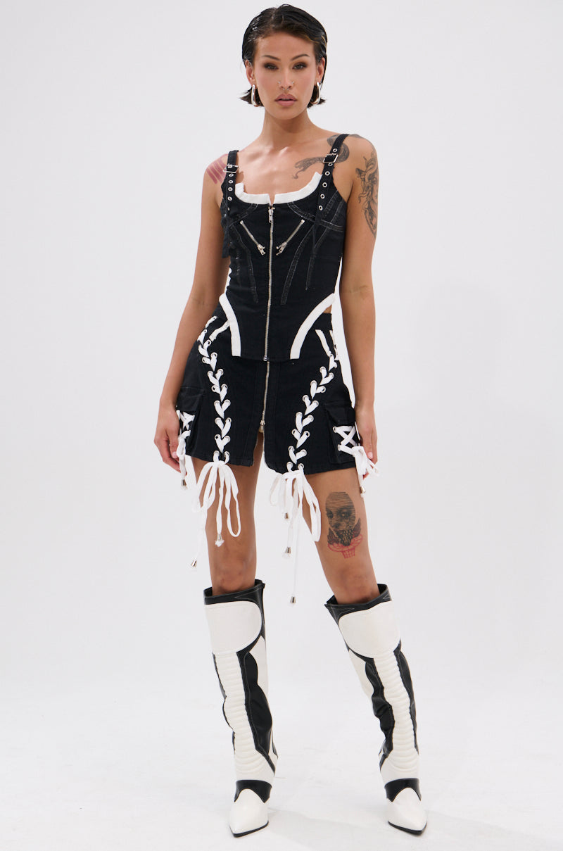 A person poses confidently in the WRAPPED UP DENIM CORSET, featuring white lacing and faux leather details, styled with zippered pockets and knee-high black and white boots. They have tattoos, slicked-back hair, and stand against a plain white background.