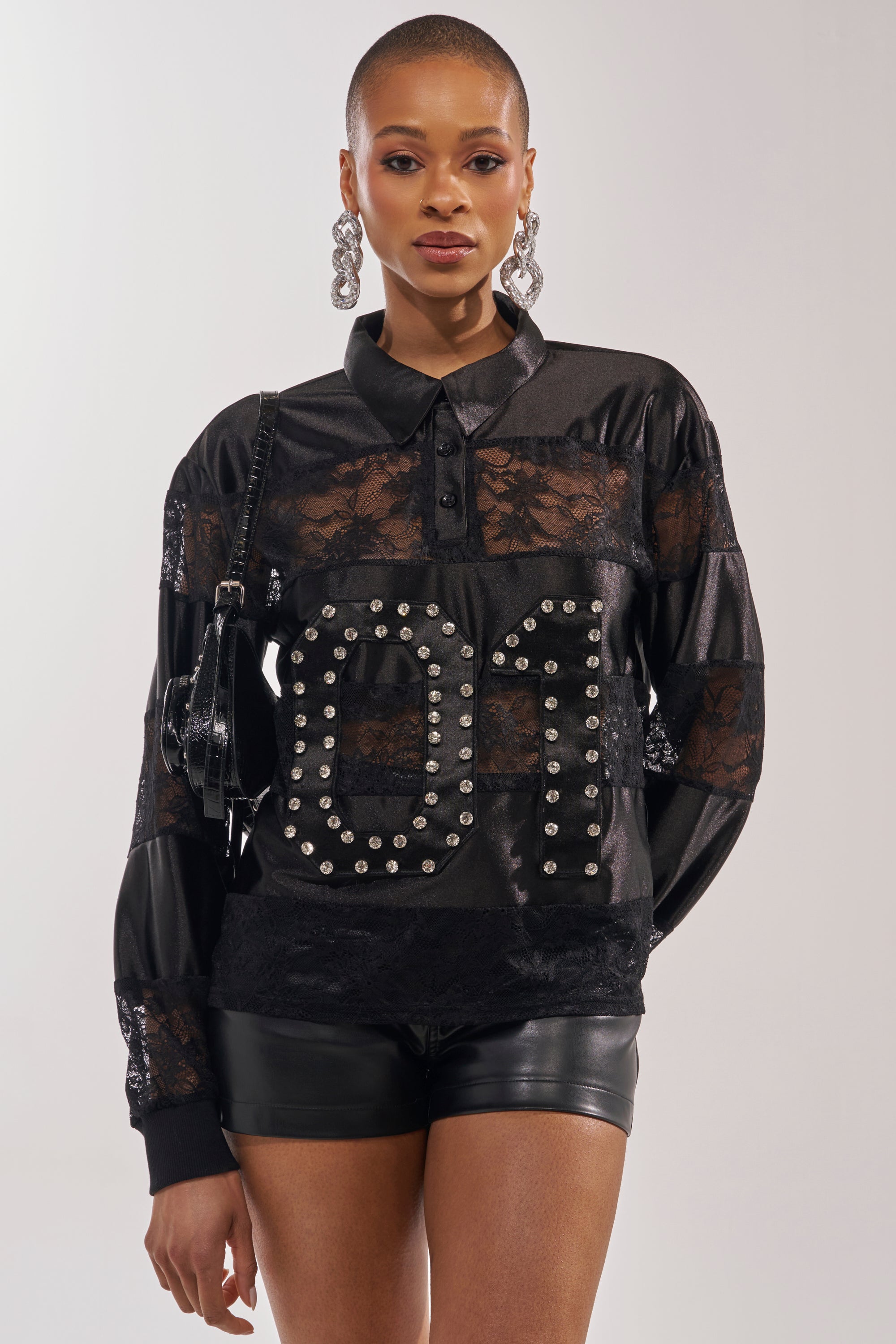 A woman with closely cropped hair wears large hoop earrings, black leather shorts, a small black purse, and the AT THE GAME PU LACE JERSEY TOP with a rhinestone "01" graphic and sheer floral lace paneling. 0