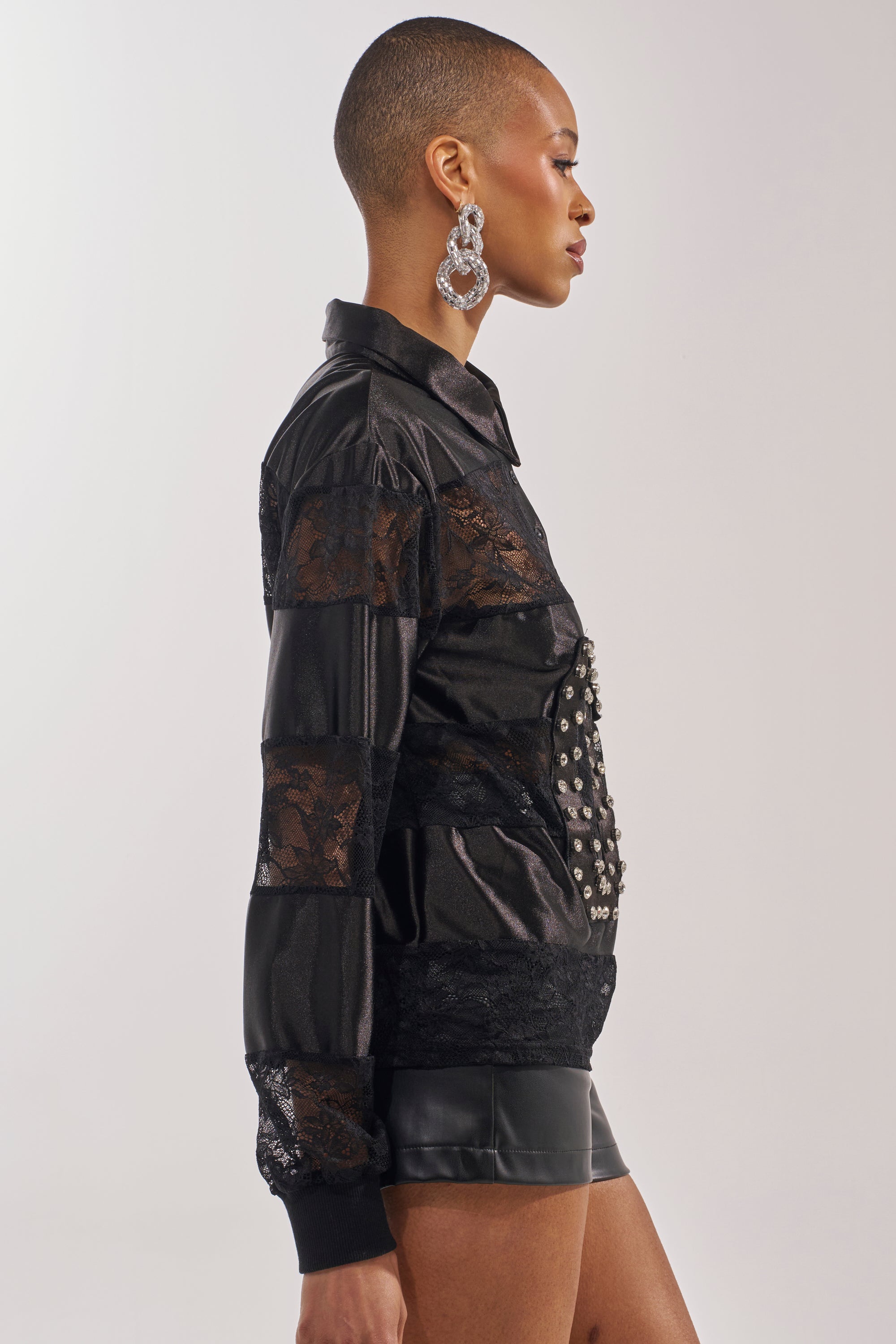 A person with a shaved head and large statement earrings wears the AT THE GAME PU LACE JERSEY TOP, featuring black lace and faux leather, paired with black leather shorts, shown in profile against a plain background. 2