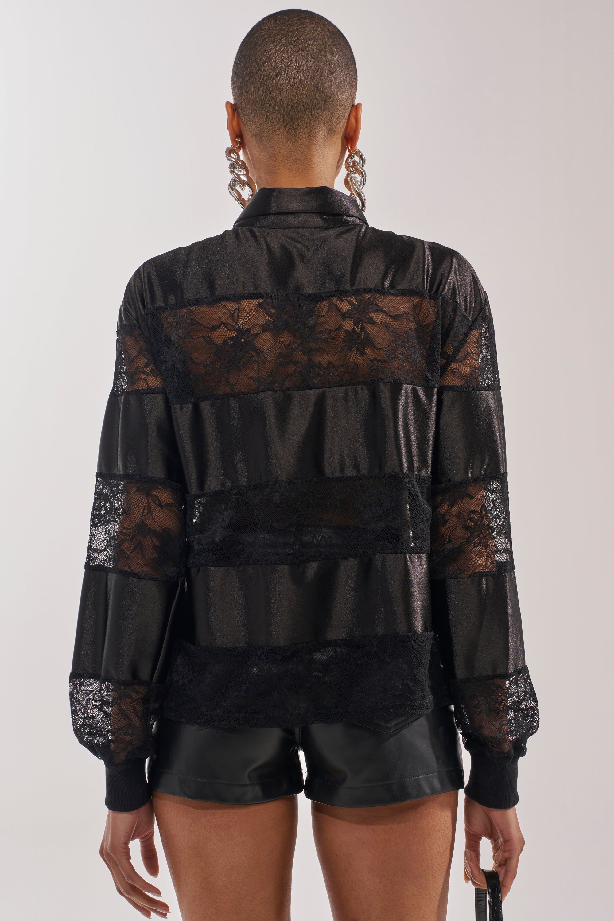 Seen from behind, a short-haired person wears the AT THE GAME PU LACE JERSEY TOP with rhinestone-studded sheer floral lace and satin long sleeves, paired with black shorts, a small bag, and large hoop earrings against a plain light background. 3