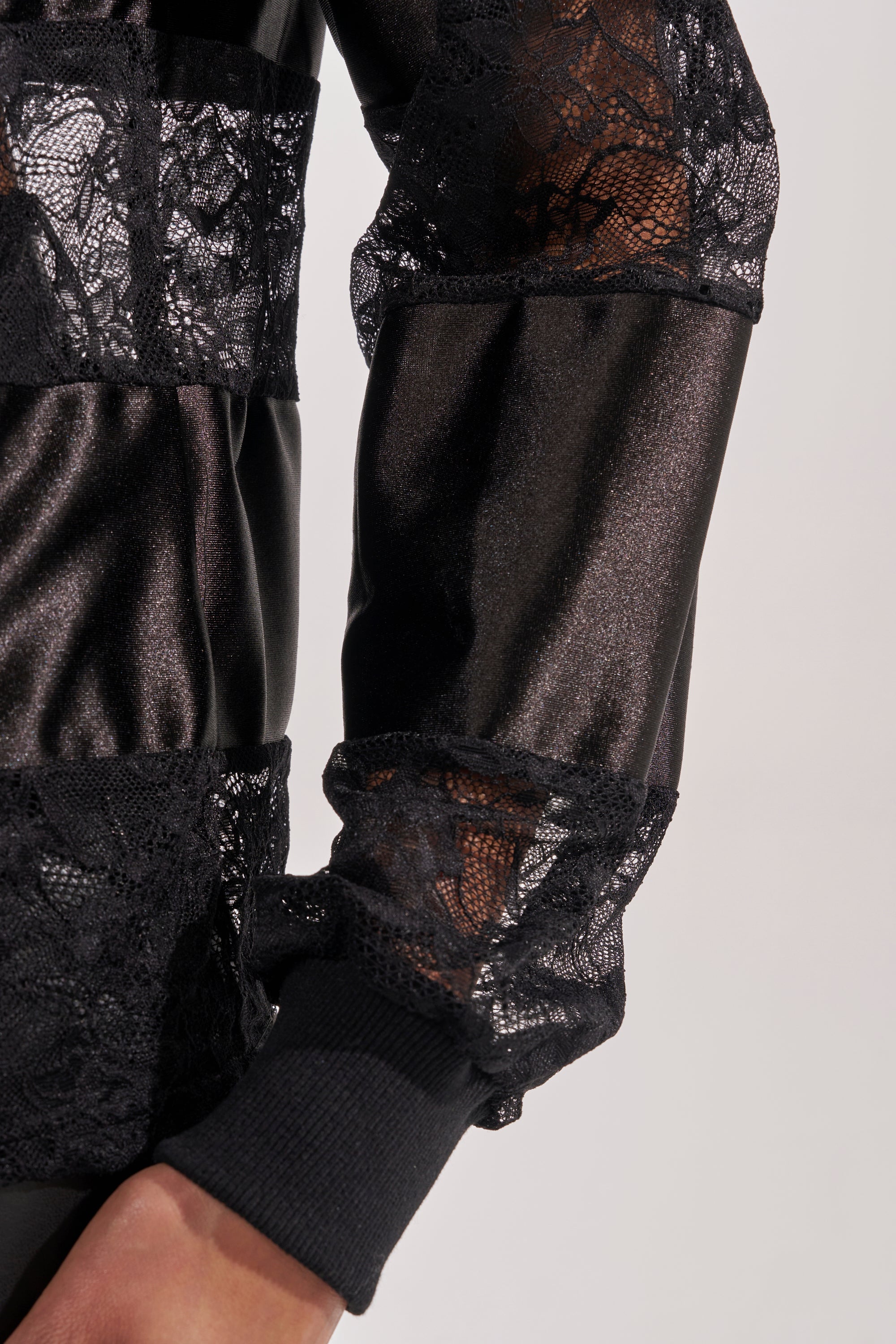 A close-up of a person's arm wearing the AT THE GAME PU LACE JERSEY TOP, which features black sheer floral lace panels, solid fabric sections, and a ribbed cuff at the wrist. 7