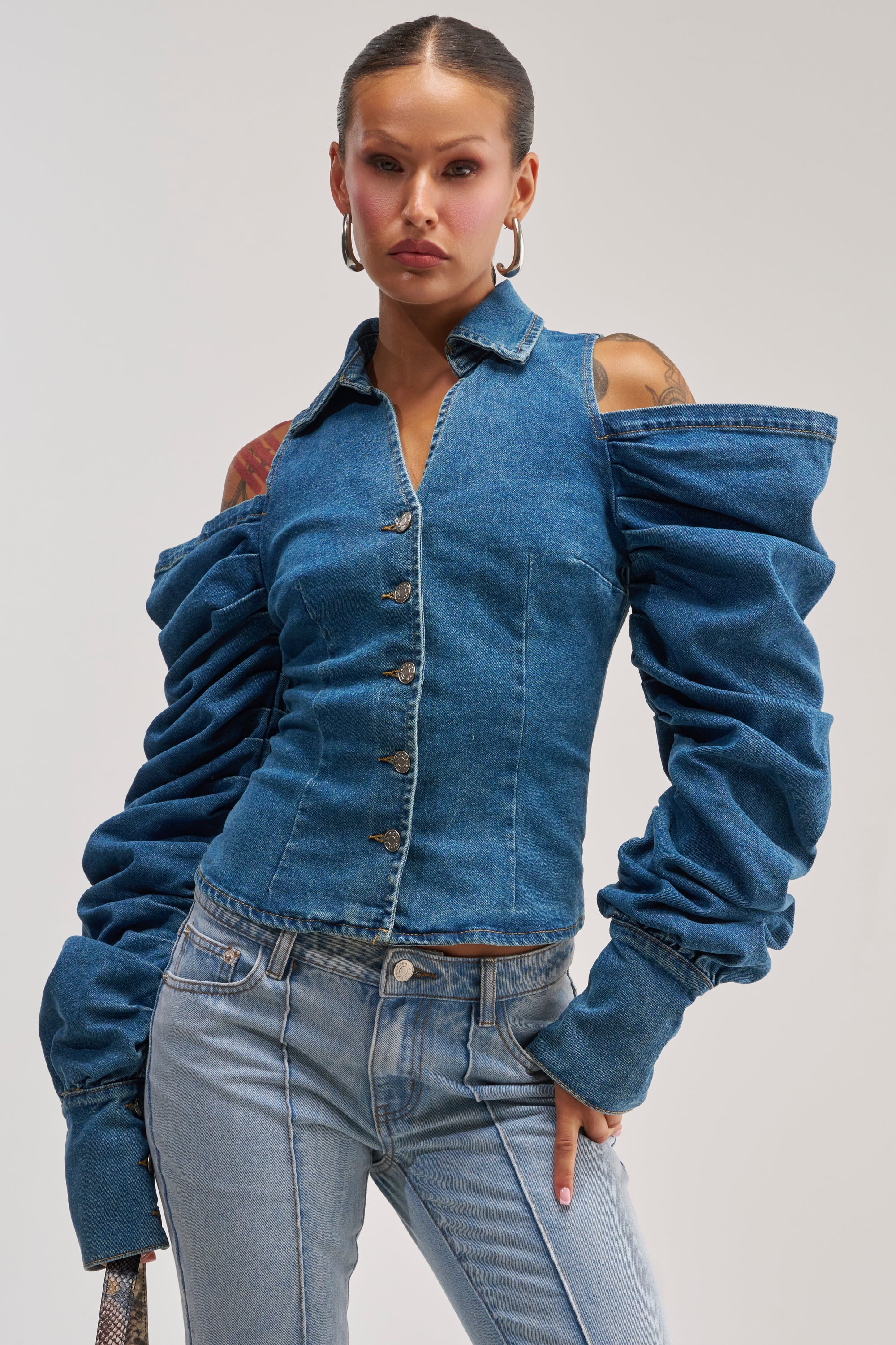 Model showcases the OFF-DUTY HEIRESS DENIM BUTTON DOWN—a striking blue denim corset top featuring off-the-shoulder puff sleeves—paired with matching jeans, confidently posing against a plain background.
