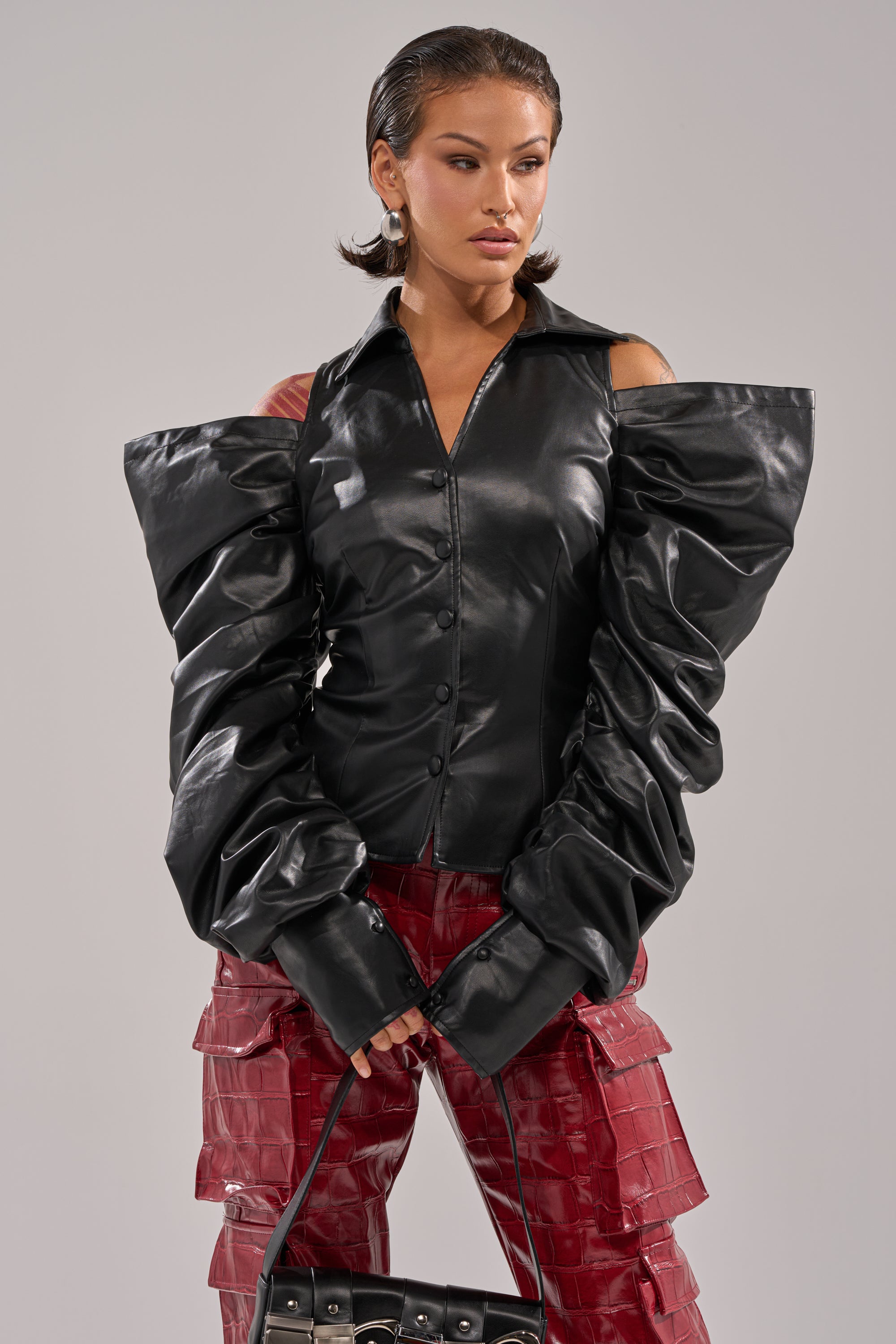 A woman wears the CRAZY IN LOVE BLACK FAUX LEATHER BUTTON DOWN BLOUSE with puffed sleeves and red textured pants, holding a black handbag as she poses confidently against a plain background. 1