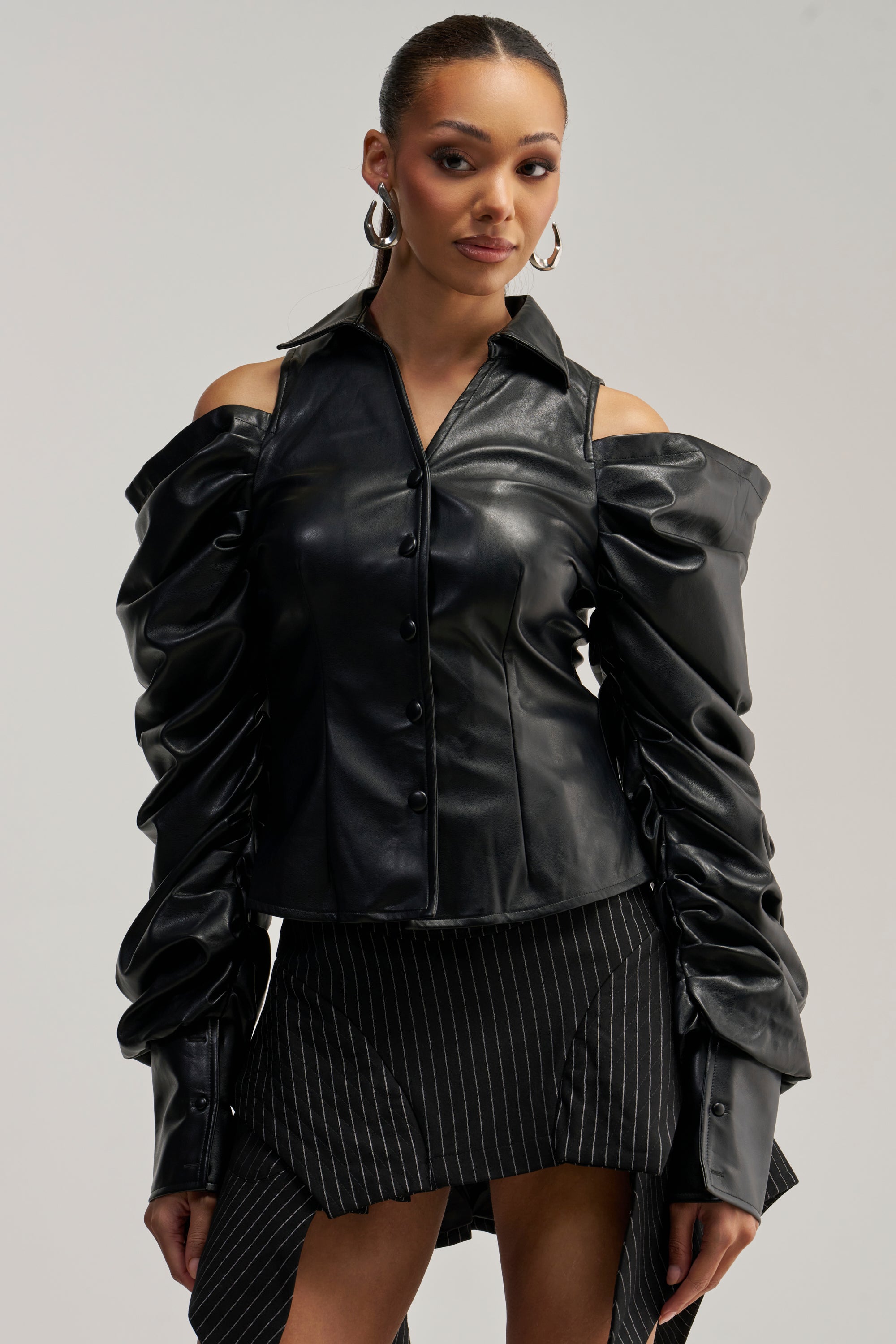 A woman wearing the CRAZY IN LOVE BLACK FAUX LEATHER BUTTON DOWN BLOUSE with cold shoulders and puffed ruched sleeves, paired with a black pinstripe asymmetrical mini skirt, stands against a plain light background.