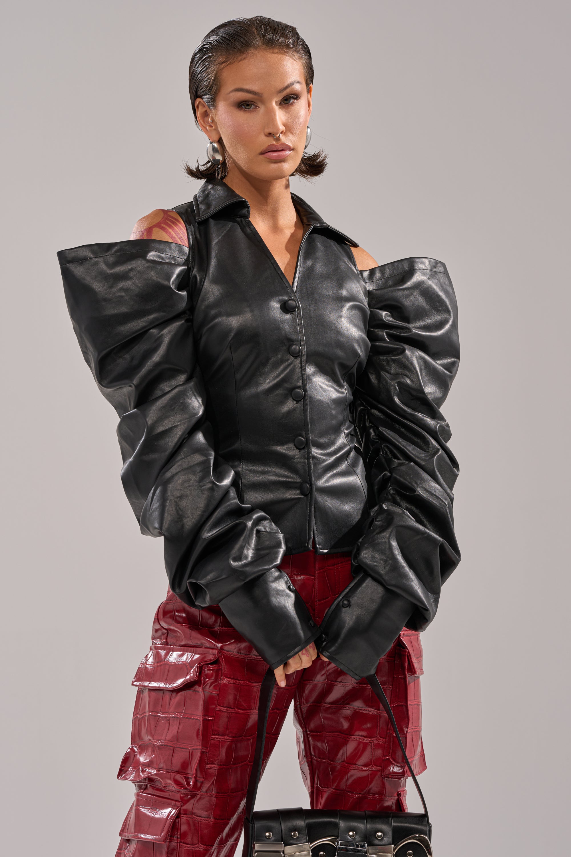 A woman with slicked-back hair models the CRAZY IN LOVE BLACK FAUX LEATHER BUTTON DOWN BLOUSE with dramatic ruched sleeves, paired with red textured pants and a small black handbag, against a neutral background. 2