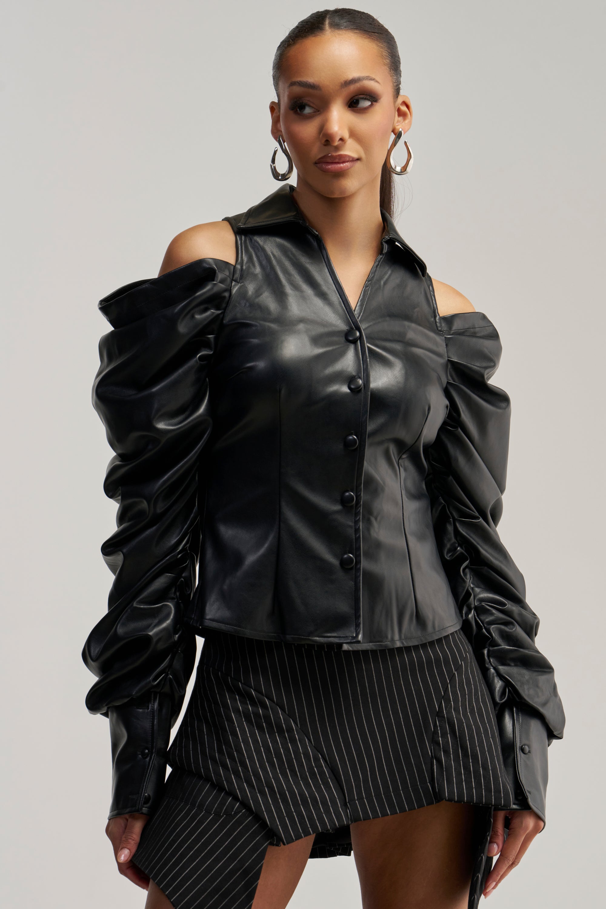 A woman wears the CRAZY IN LOVE BLACK FAUX LEATHER BUTTON DOWN BLOUSE styled off the shoulder, paired with a pinstripe skirt featuring an asymmetric hem for an edgy streetwear look. She accessorizes with hoop earrings and has her hair slicked back.