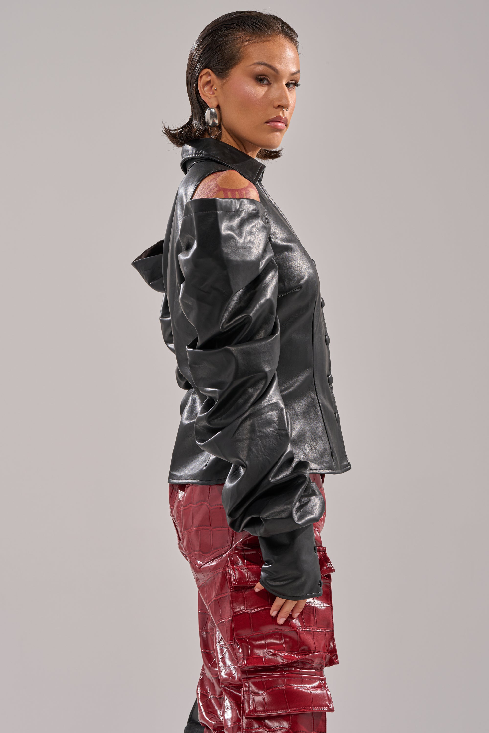 A woman with slicked-back hair poses sideways in the CRAZY IN LOVE BLACK FAUX LEATHER BUTTON DOWN BLOUSE, featuring ruched sleeves, paired with shiny red textured pants against a plain background. 3