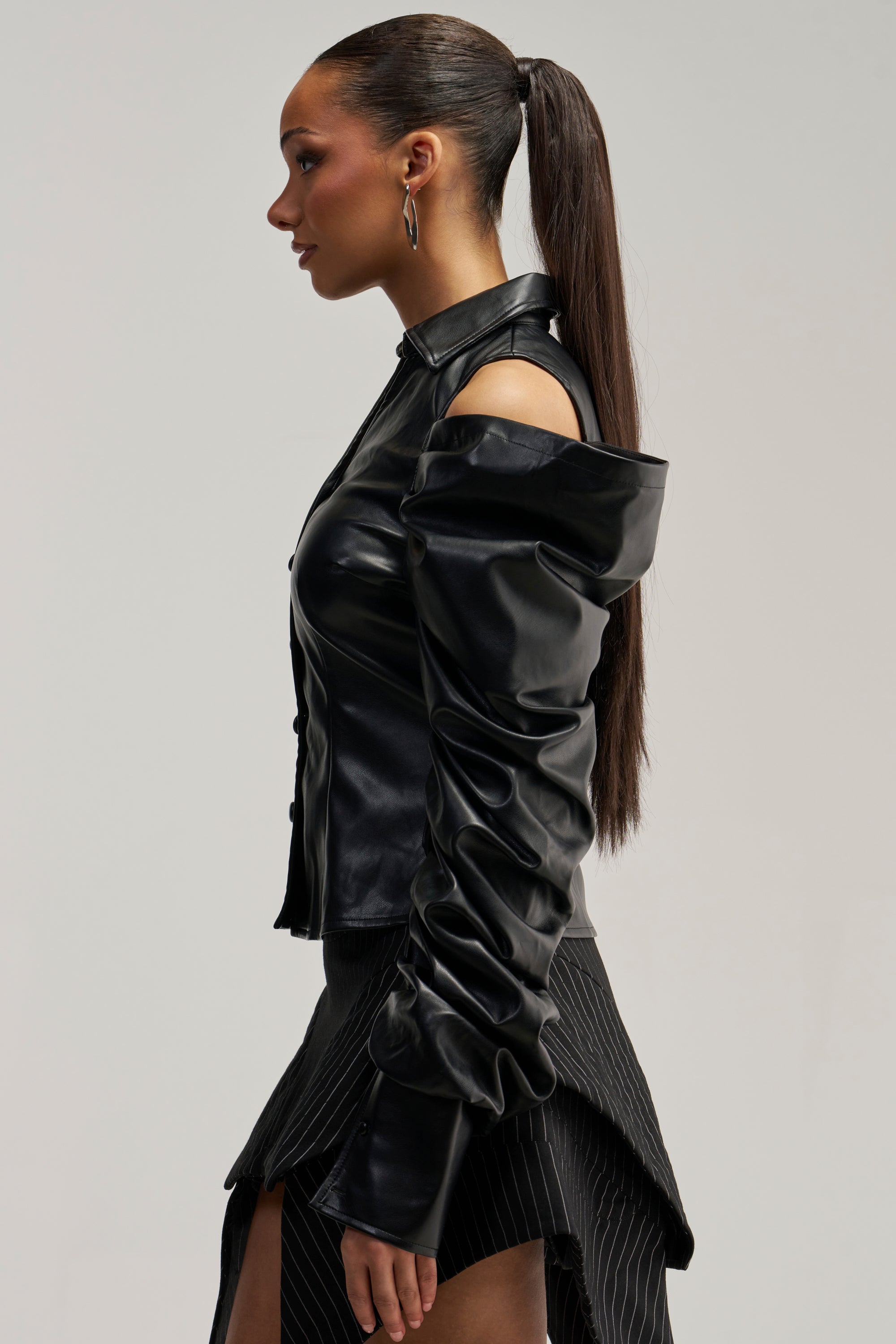Wearing the CRAZY IN LOVE BLACK FAUX LEATHER BUTTON DOWN BLOUSE with exposed shoulders and long ruched sleeves, she pairs it with a black pinstripe skirt featuring a thigh-high slit for a bold streetwear look. Her hair is styled in a long, sleek ponytail.
