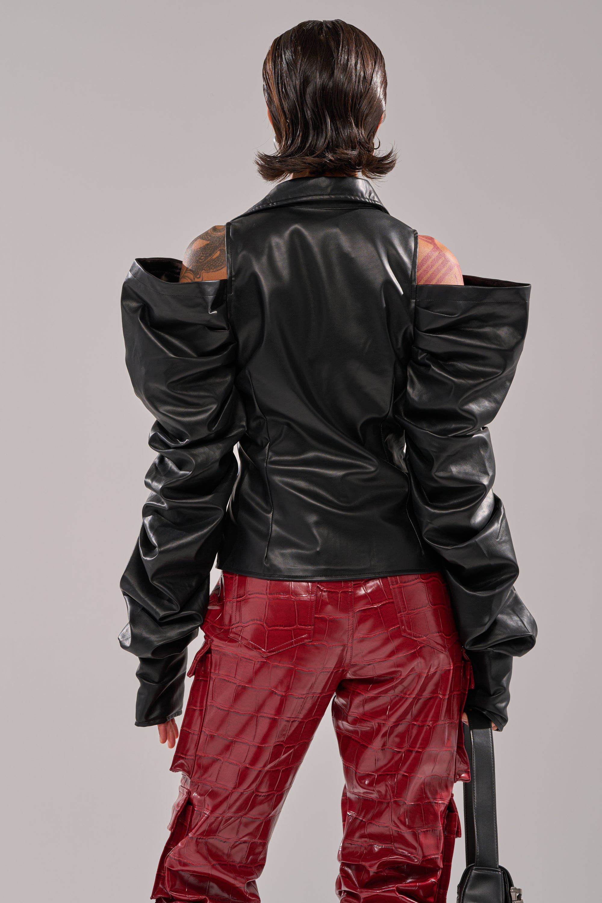 A person stands facing away, wearing the CRAZY IN LOVE BLACK FAUX LEATHER BUTTON DOWN BLOUSE with dramatic ruched sleeves and red, shiny textured pants. Their short hair is slicked back, and they hold a black bag by their side. 4